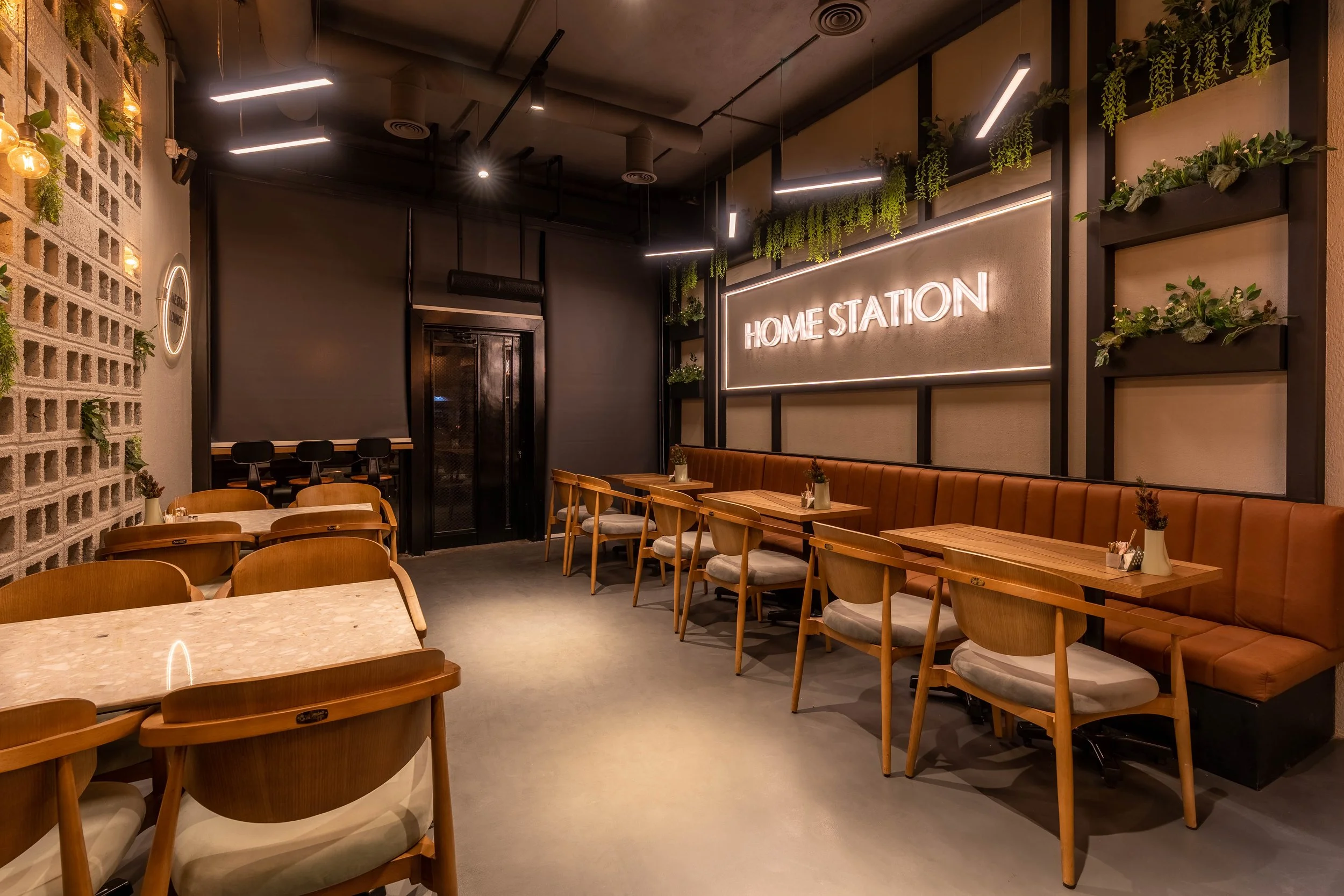 Empty modern cafe interior with wooden chairs and tables, a brown cushioned bench along the wall, decorative plants, and a neon sign that says "HOME STATION"