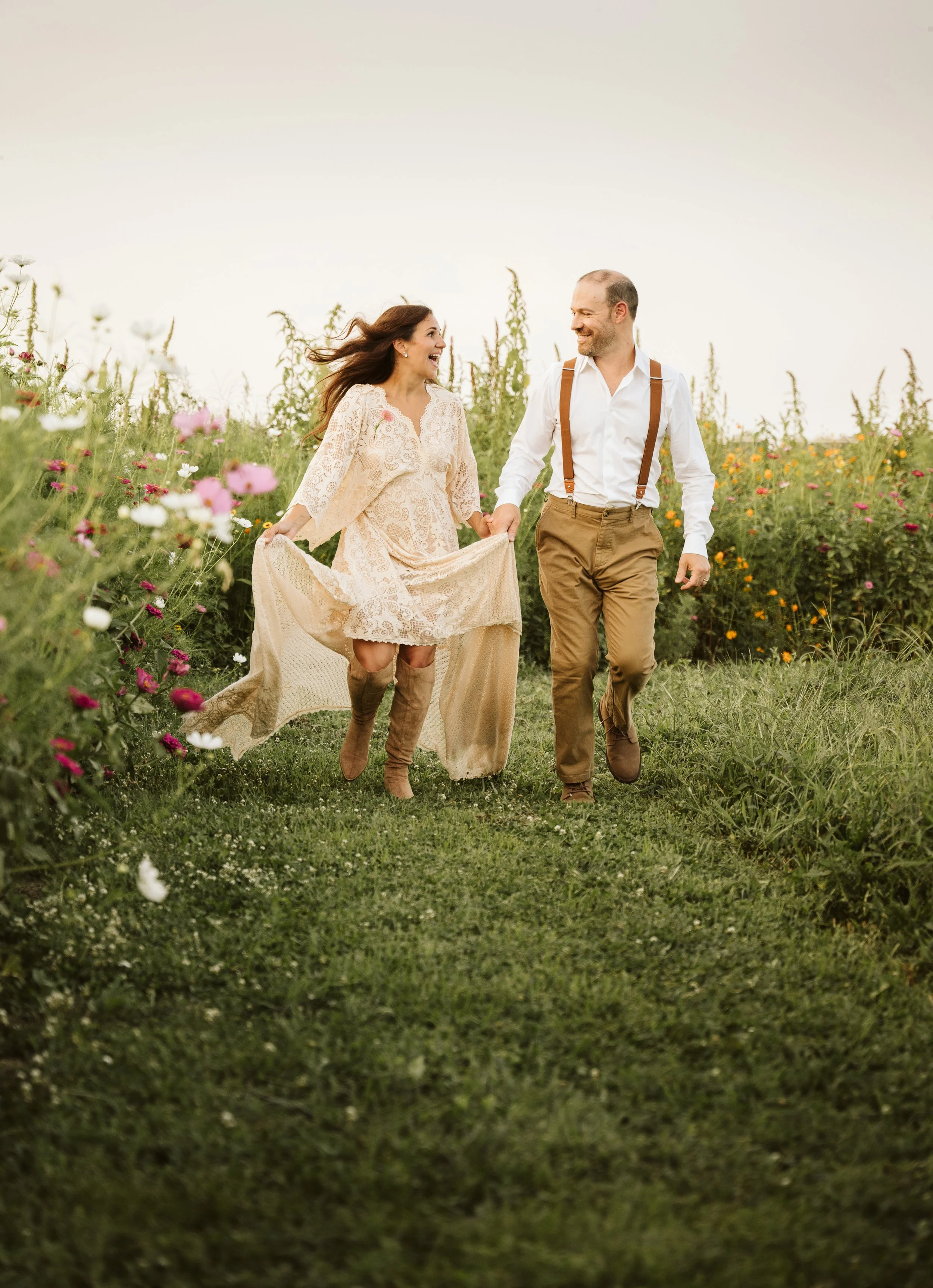 A smiling couple walking hand in hand through a field of flowers at sunset, with the woman wearing a lace dress and boots and the man in dress pants with suspenders.