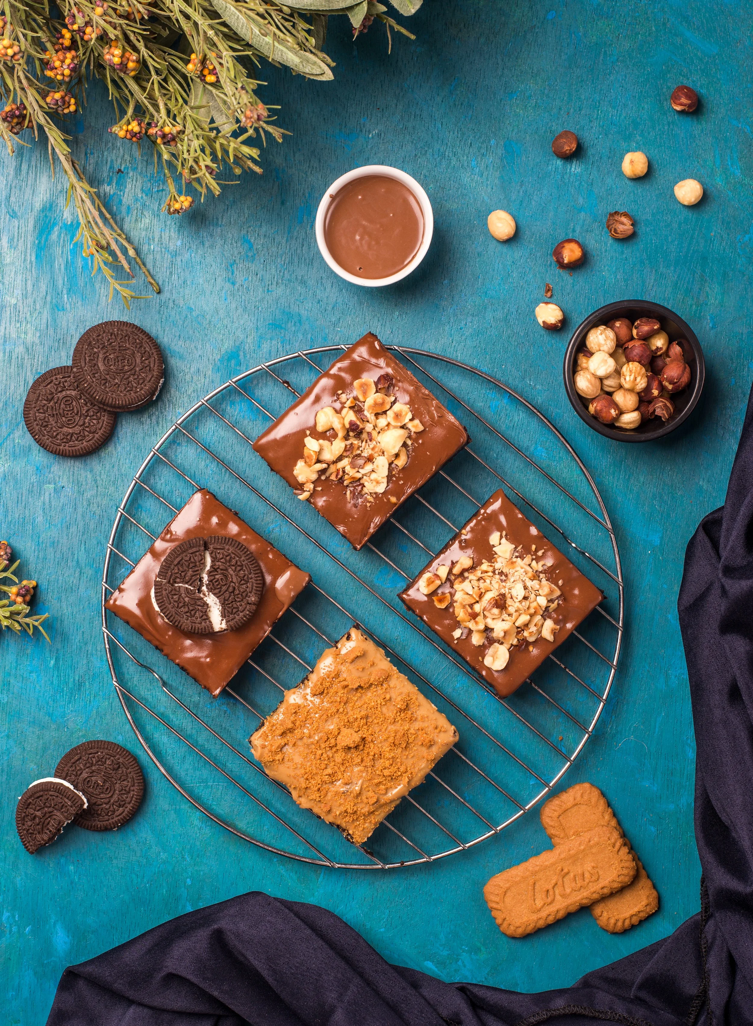 Assorted plated dessert brownies topped with chopped nuts, surrounded by chocolate cookies, a small cup of chocolate sauce, a bowl of hazelnuts, and some cookies on a vibrant blue background.