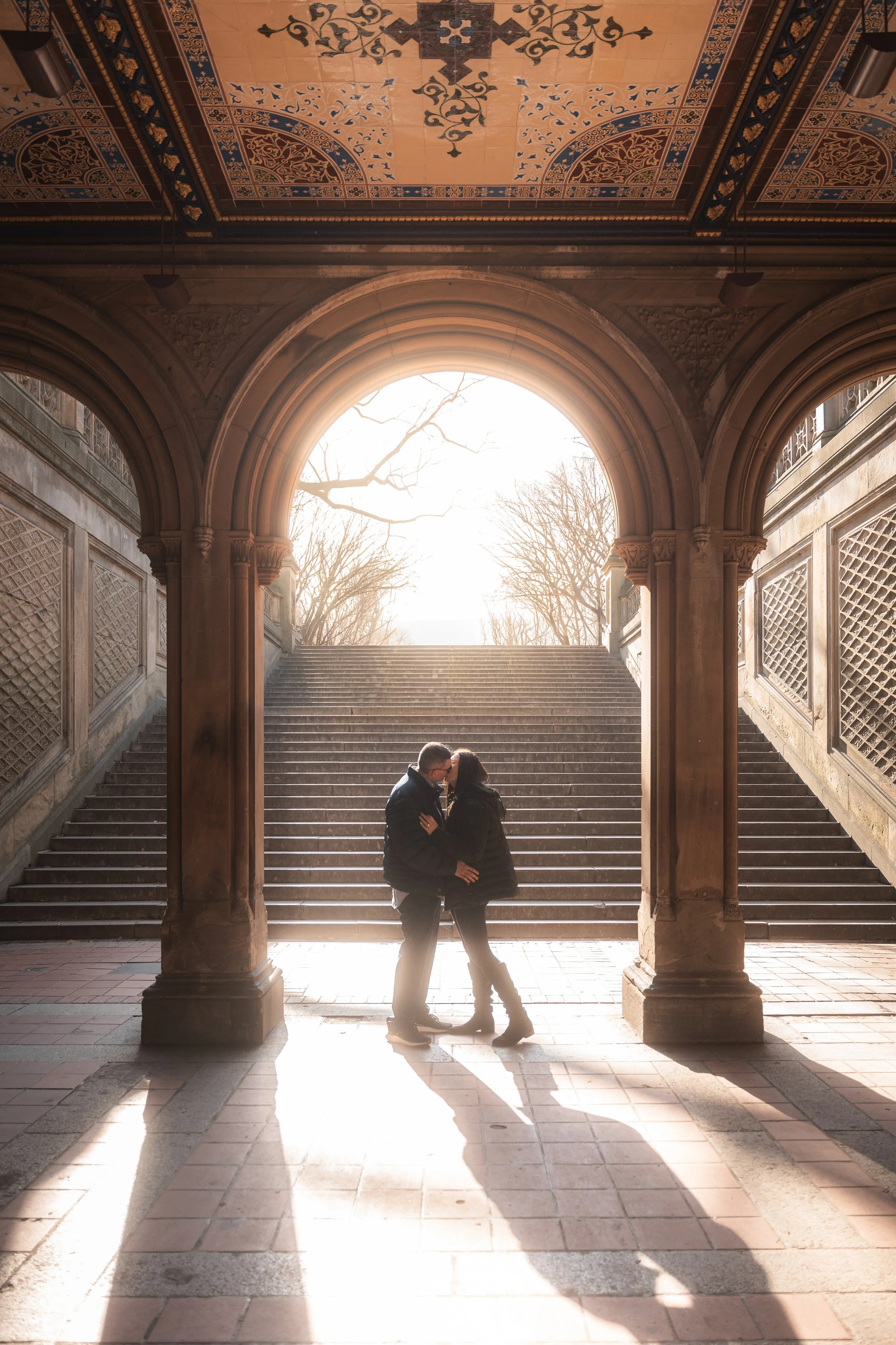 A couple is kissing under an ornate archway in Central Park NYC with stairs and trees in the background during sunset.