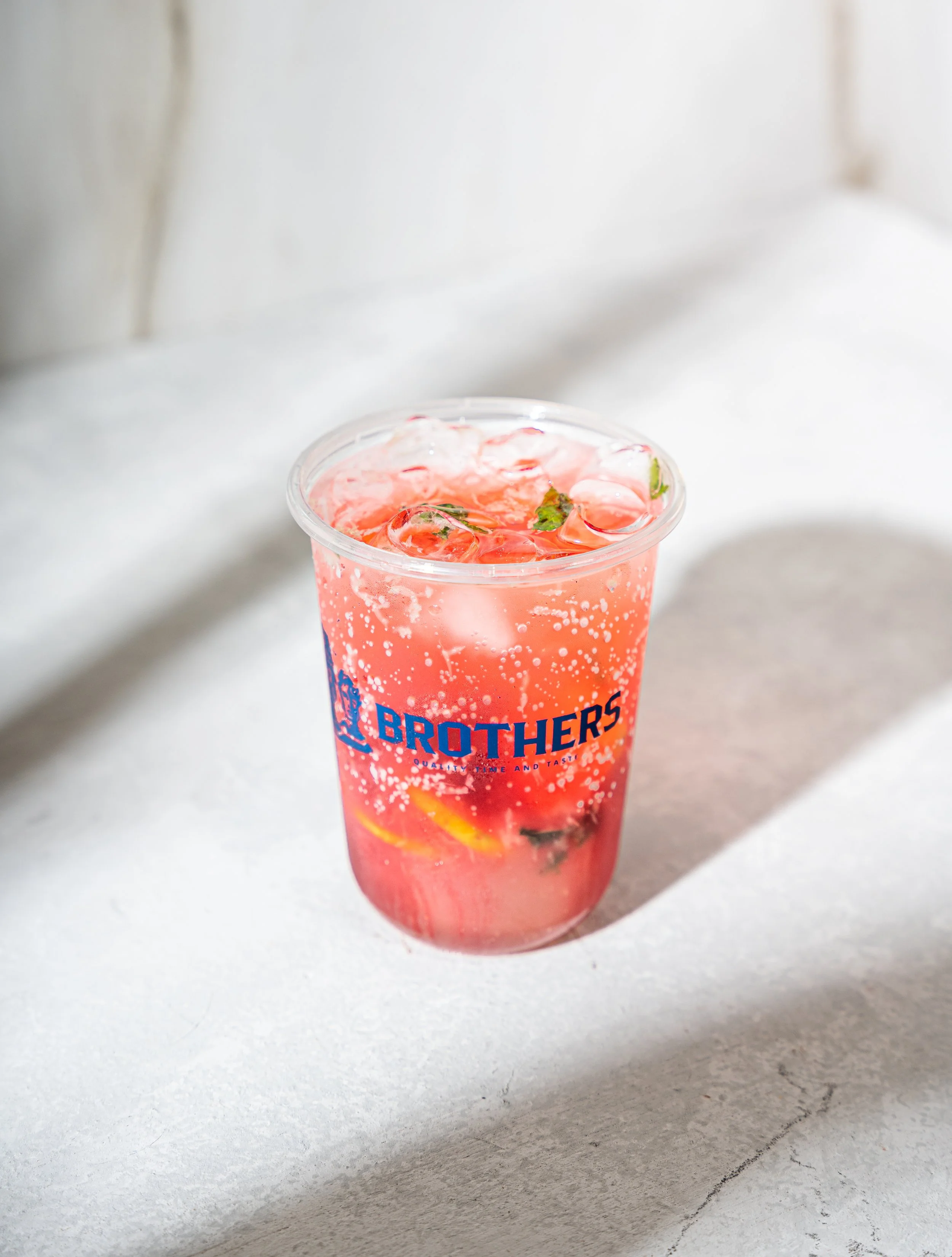 Pink soda in a clear plastic cup with ice, lemon, and mint, with 'BROTHERS' printed on the cup, on a white surface with sunlight.