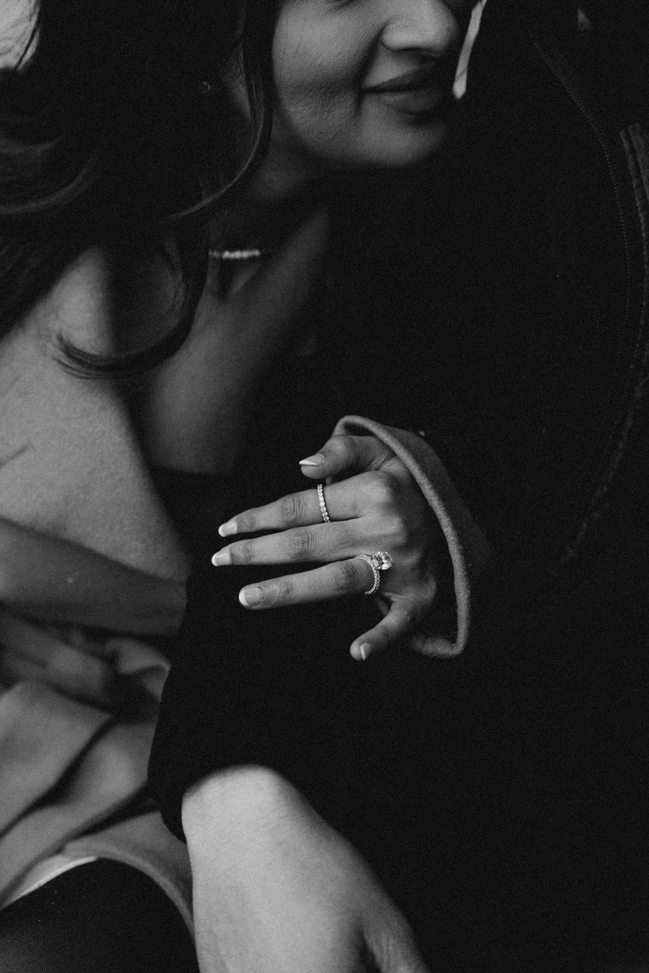 A black and white close-up photo of two women embracing, with one resting her head on the other's shoulder. The visible woman's face has a gentle smile, and their hands, adorned with rings, are clasped together.