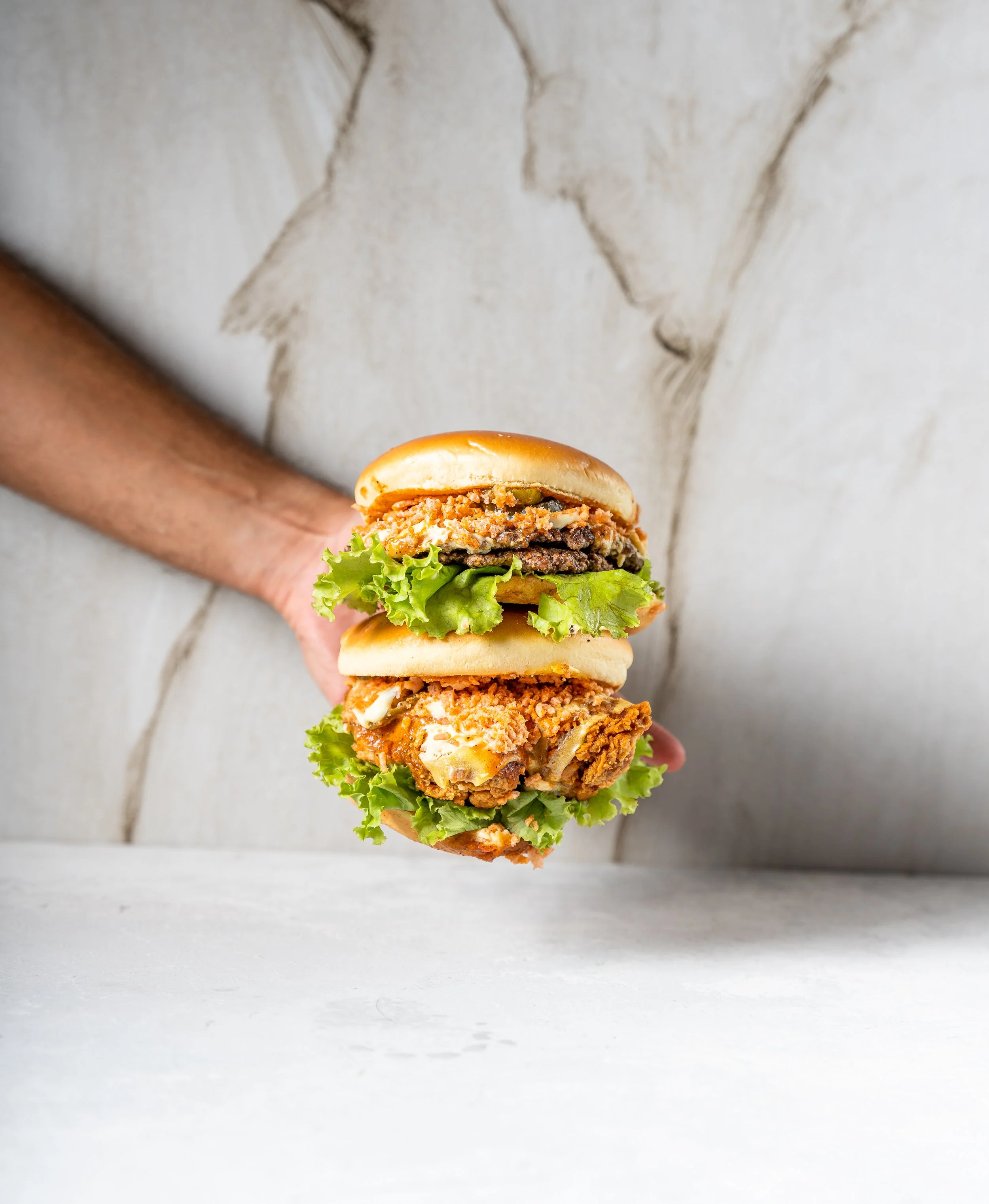 Person holding two burgers with lettuce, fried chicken, and beef patties against a light background.