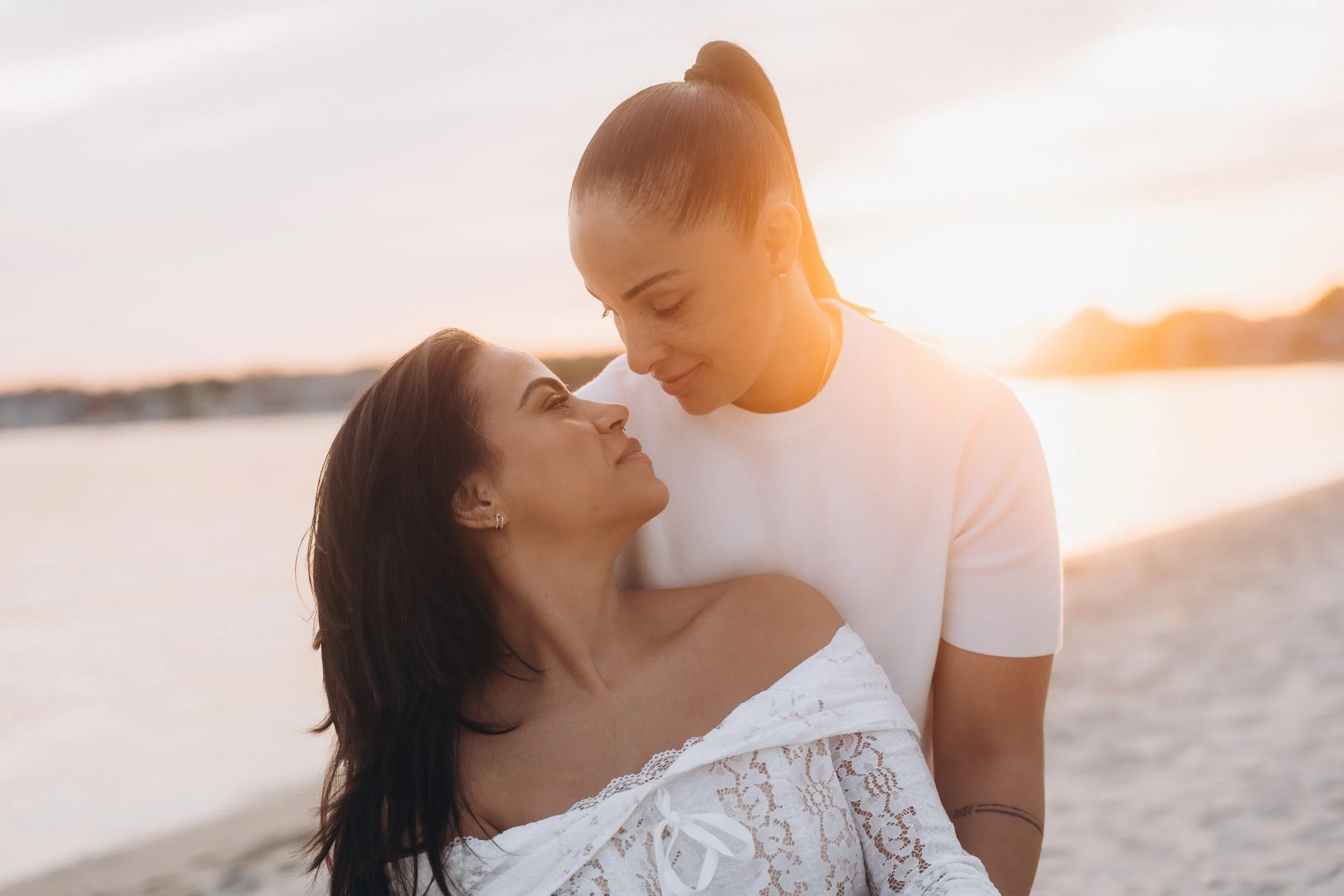 A couple standing close together on a beach at sunset, looking into each other's eyes with the ocean in the background.