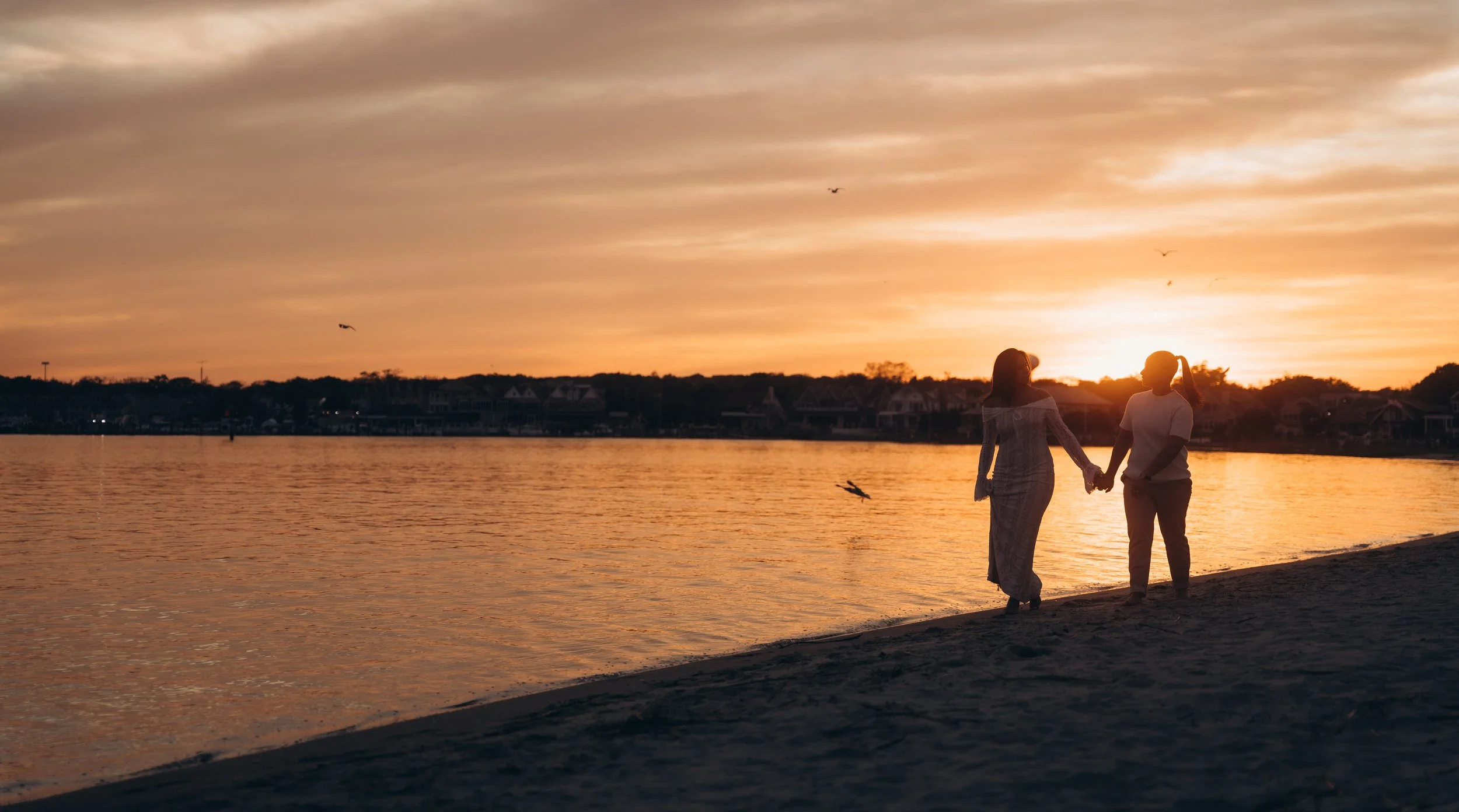 Two women holding hands and walking along a beach at sunset with a calm body of water and houses on the distant shoreline.