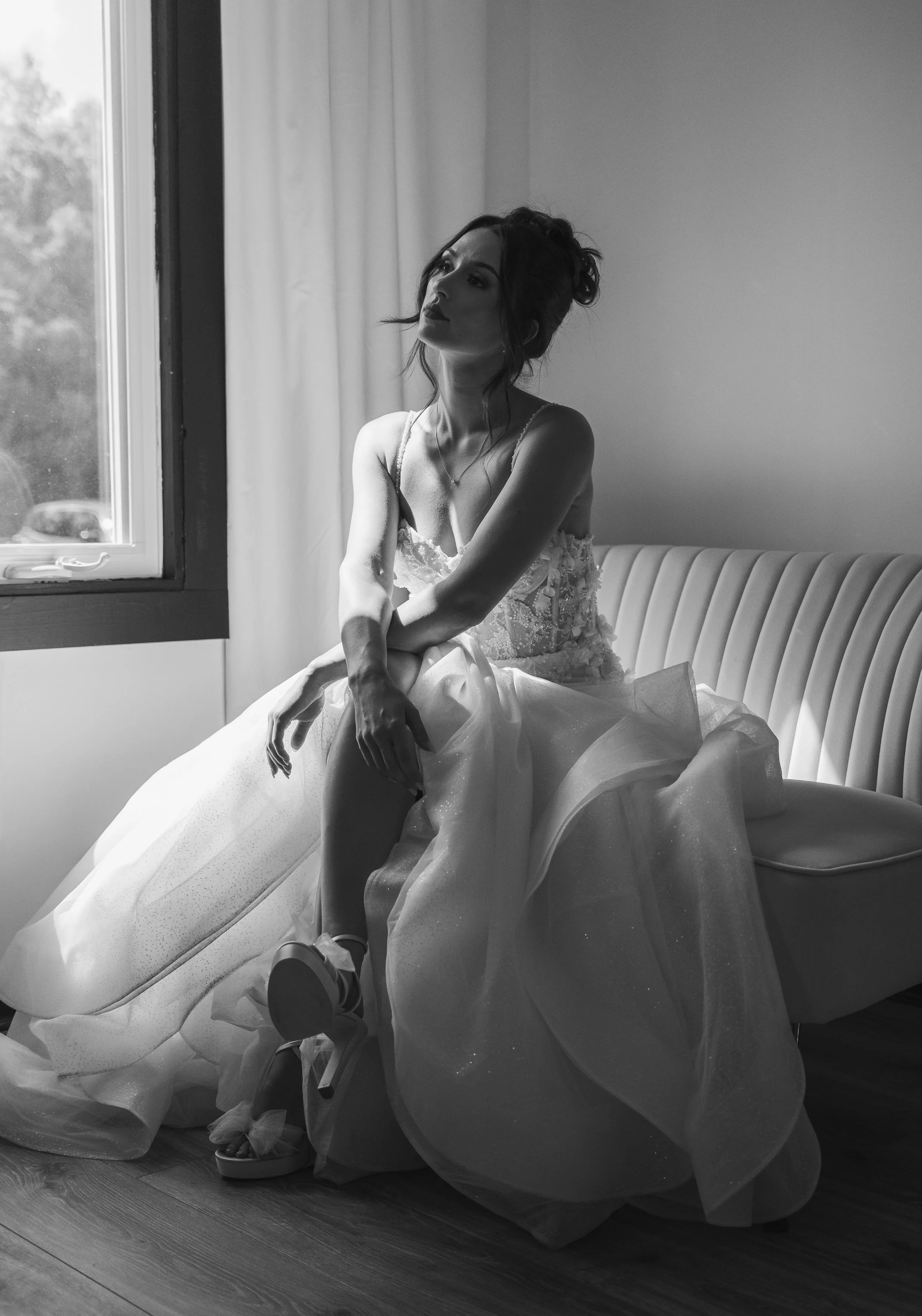 A woman in a wedding dress sitting on a couch near a window, looking contemplative in black and white.