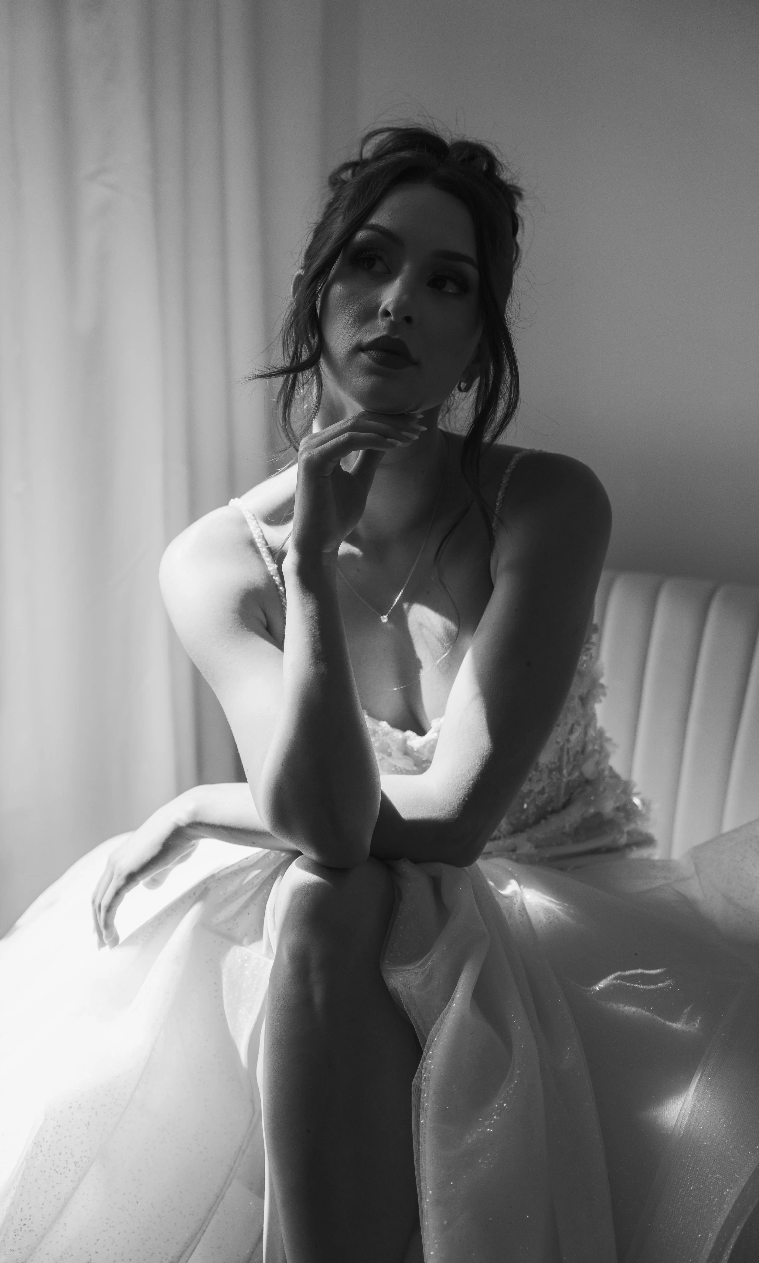 A woman in a wedding dress sitting on a bed, with her chin resting on her hand, in a black-and-white photograph.