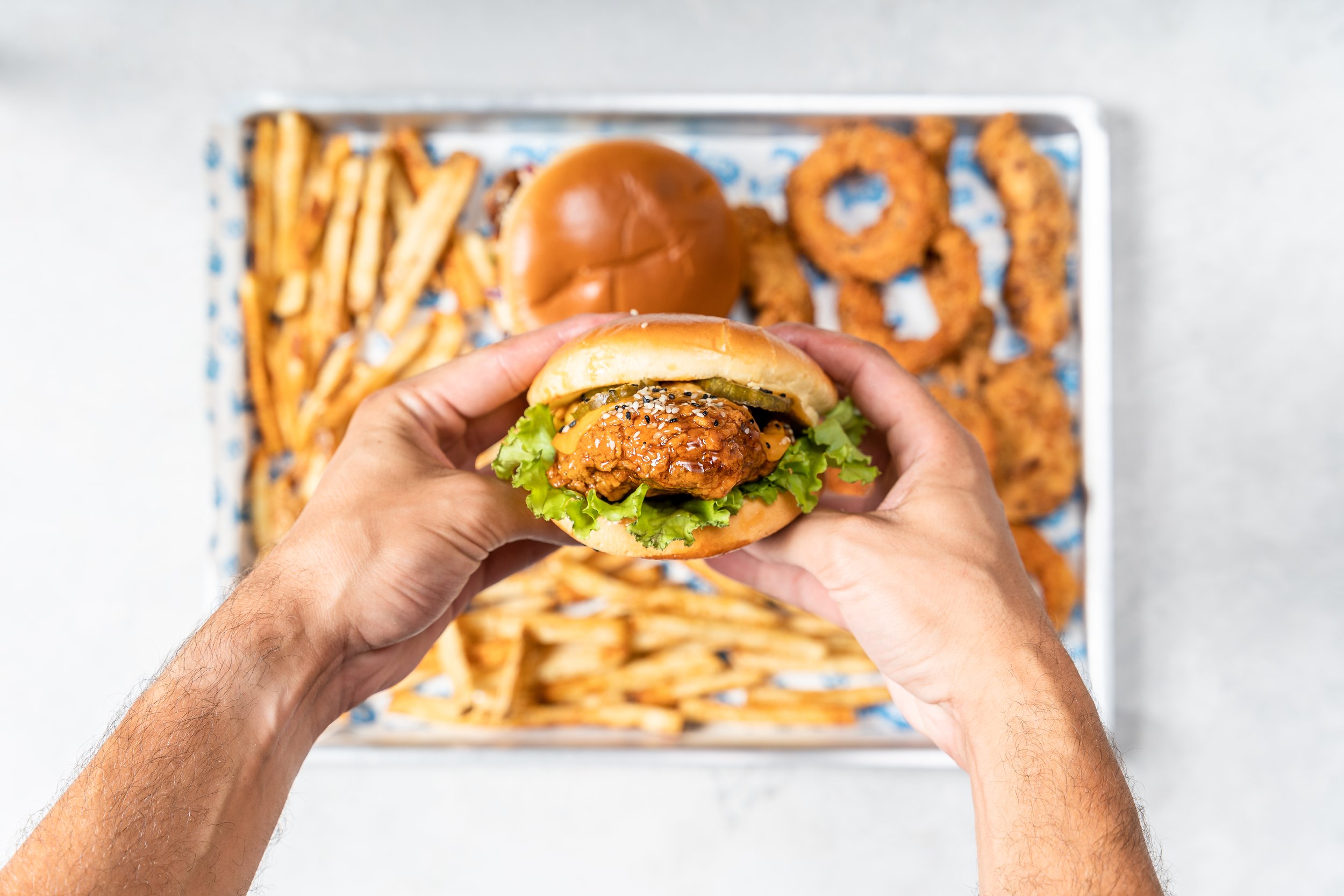 Person holding a fried chicken sandwich with lettuce, pickles, and sauce, with a tray of fries, onion rings, and a burger in the background.