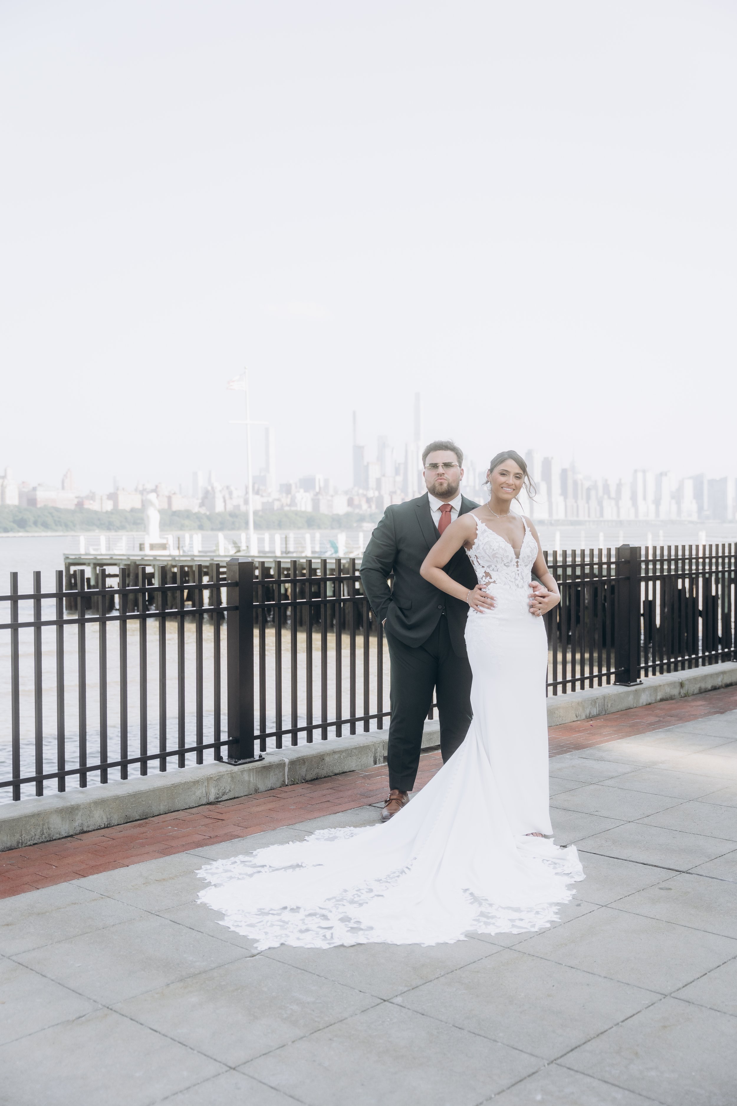 A bride and groom posing outdoors on a city waterfront with a skyline in the background. The bride is in a white lace wedding gown with a train, and the groom is in a black suit with a red tie.