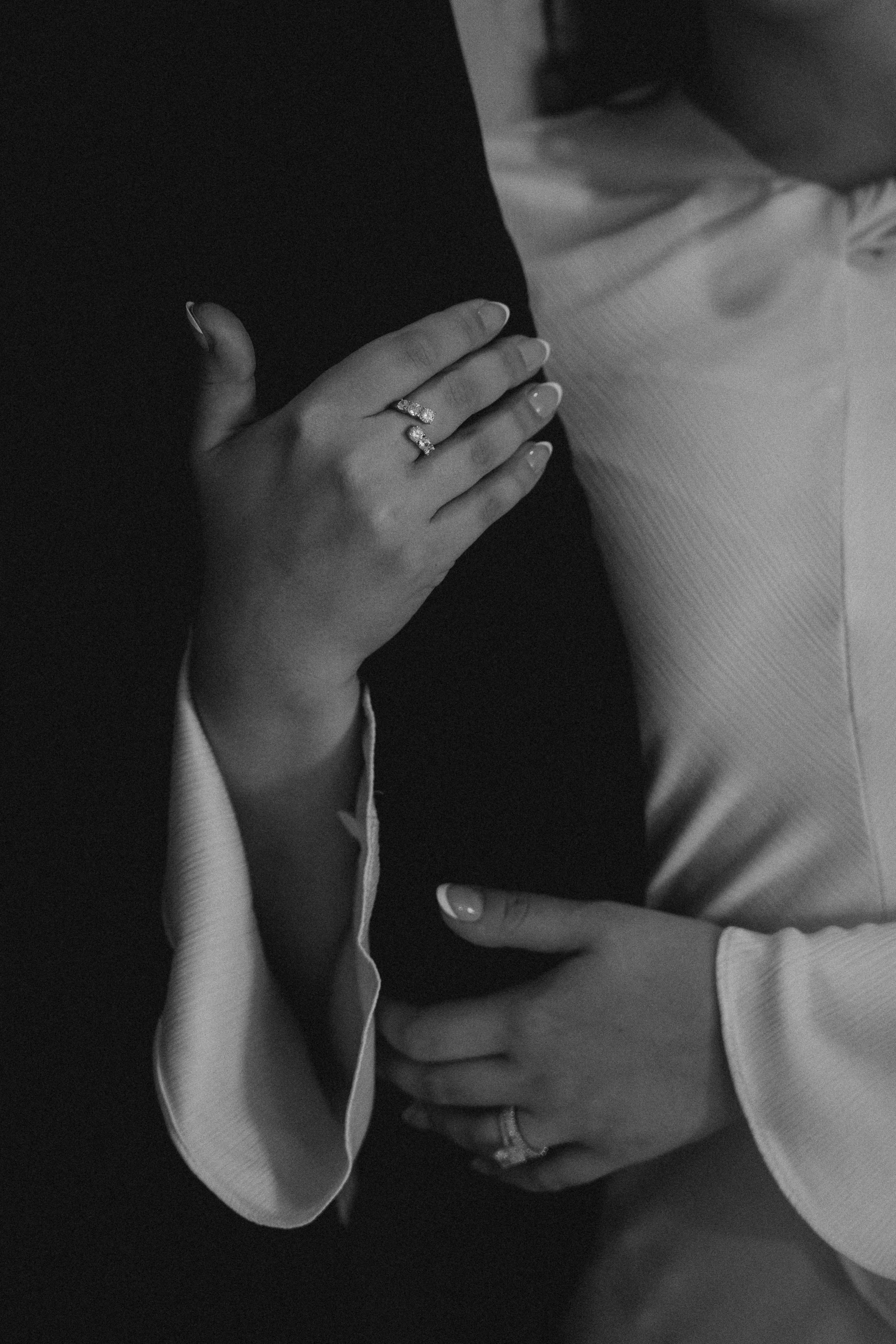 Black and white photo of two people, showing only parts of their bodies. One person’s hand is resting on the other’s chest, both wearing rings.