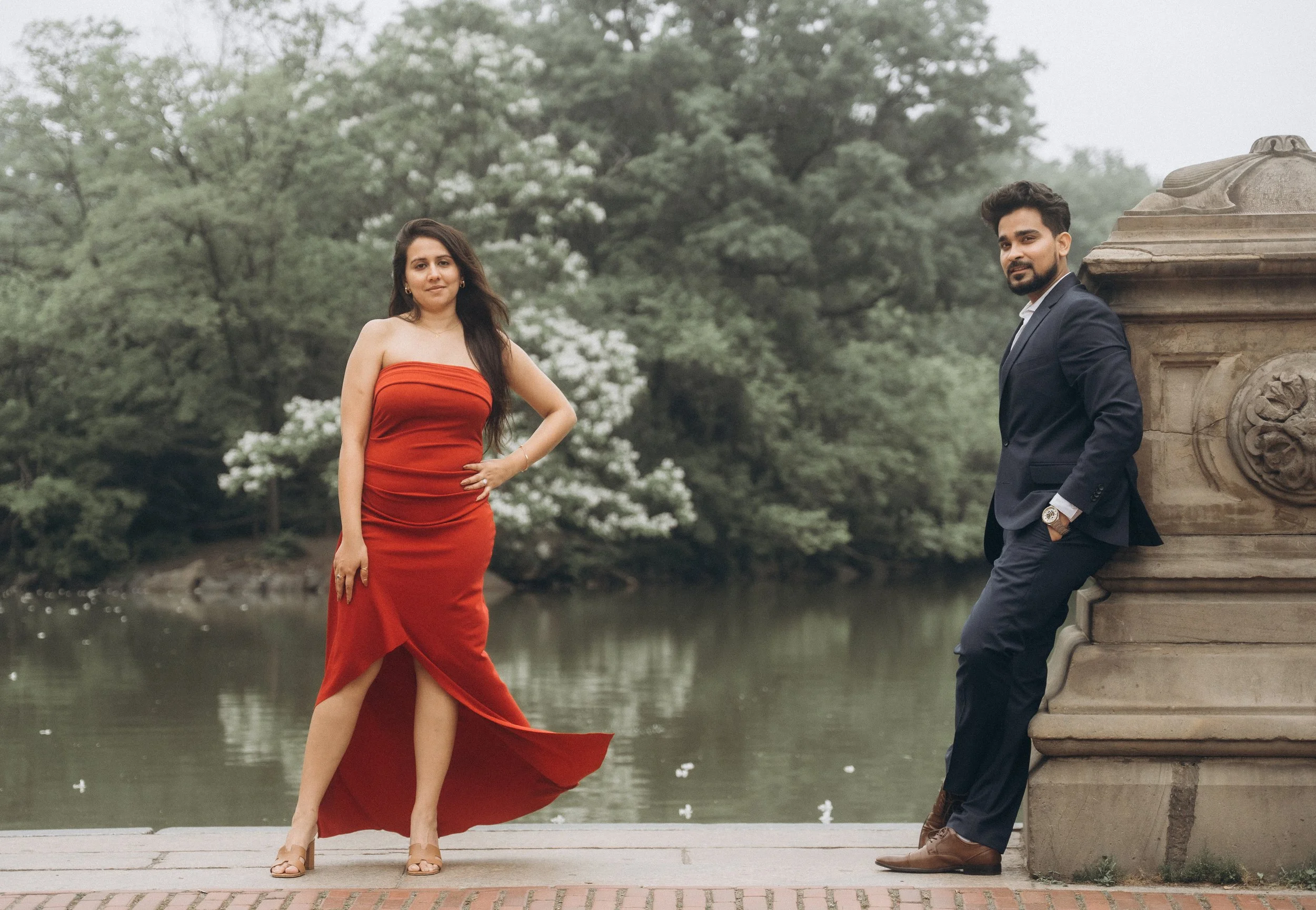 A woman in a strapless red dress and high heels stands on a paved area near a lake in Central Park NYC, with trees and white blossoms in the background. A man in a navy suit leans against a stone structure nearby.