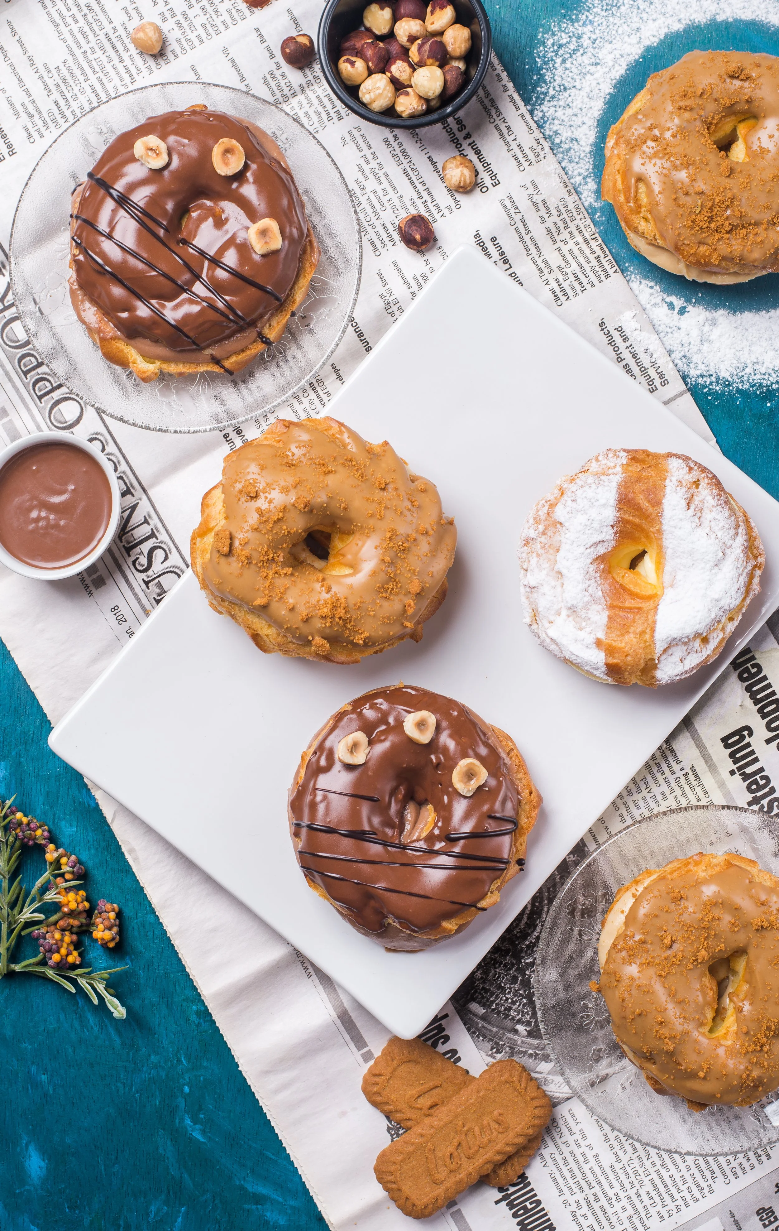 Assorted donuts with various toppings on a white plate and glasses, a small bowl of hazelnuts, a chocolate sauce cup, and cookies on a blue table covered with newspaper