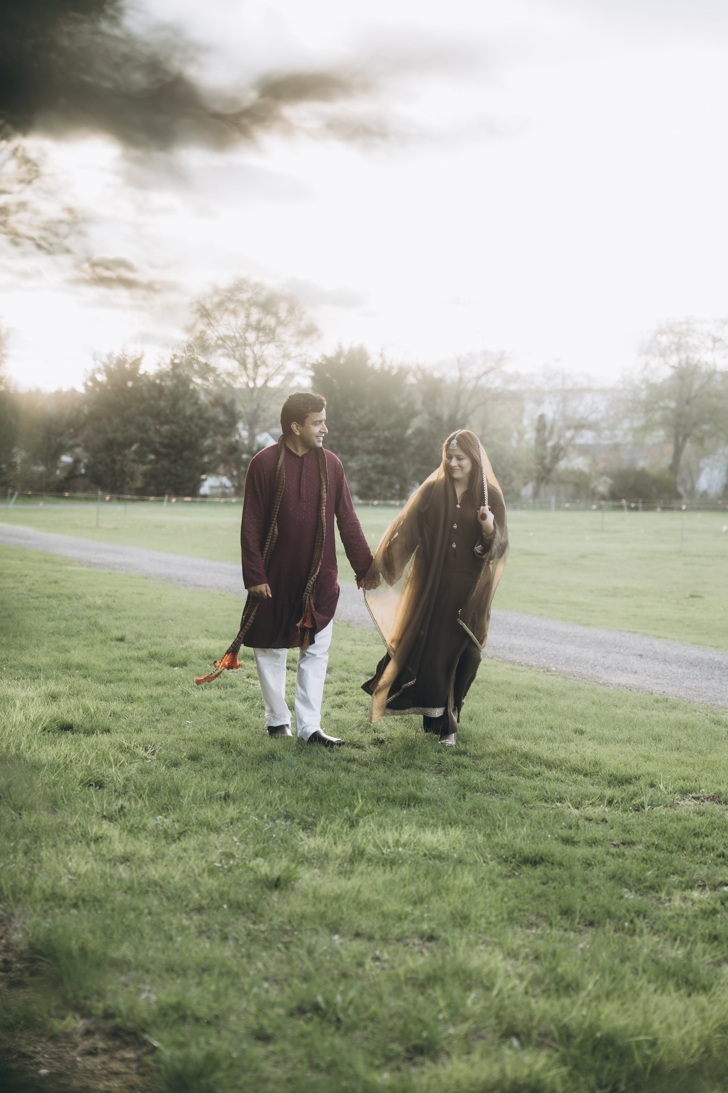 A smiling couple walking hand-in-hand on a grassy field during daytime, dressed in traditional South Asian attire, with trees and cloudy sky in the background.