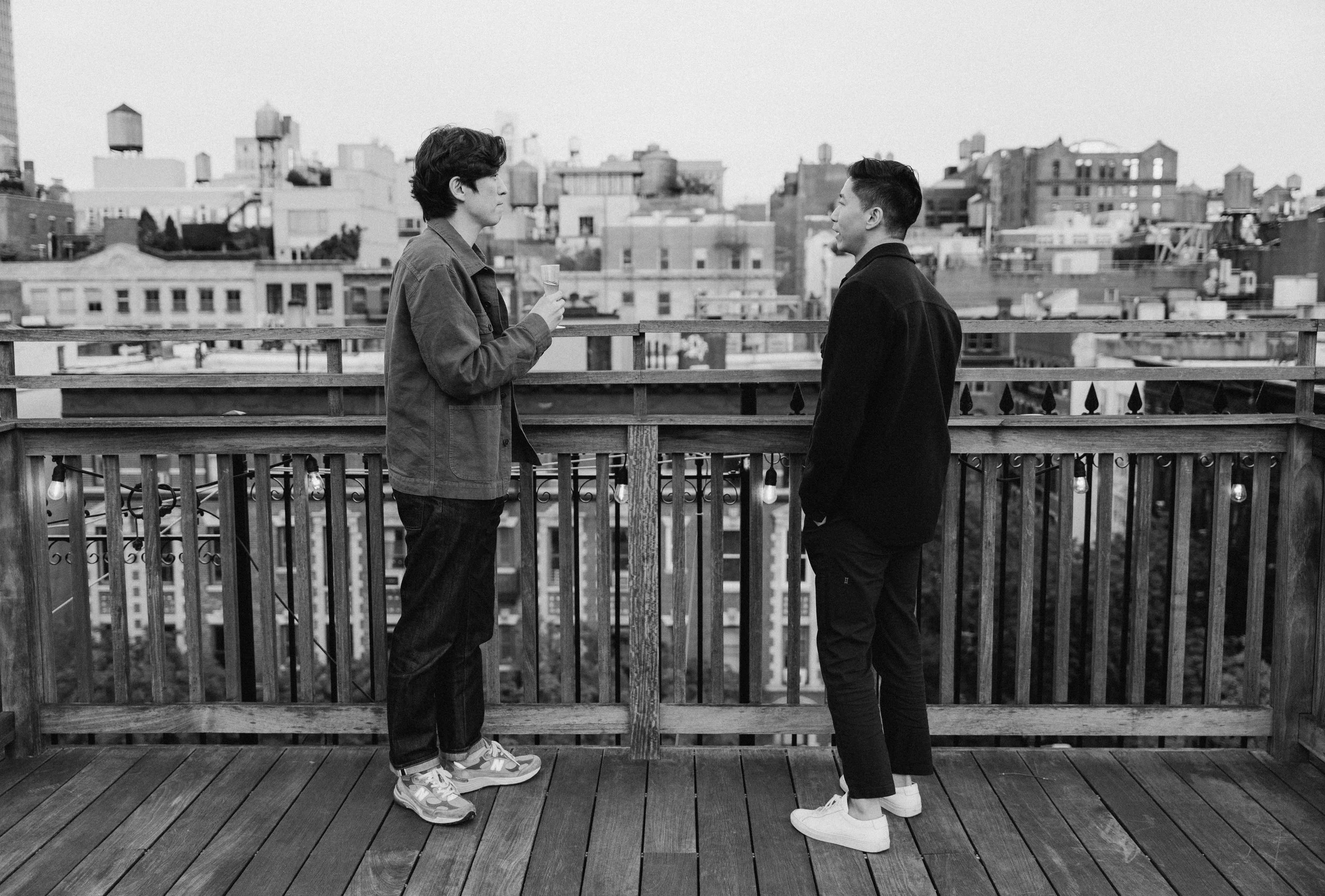 Two young men standing on a rooftop deck engaged in conversation, with one holding a drink, in front of city skyline in black and white.