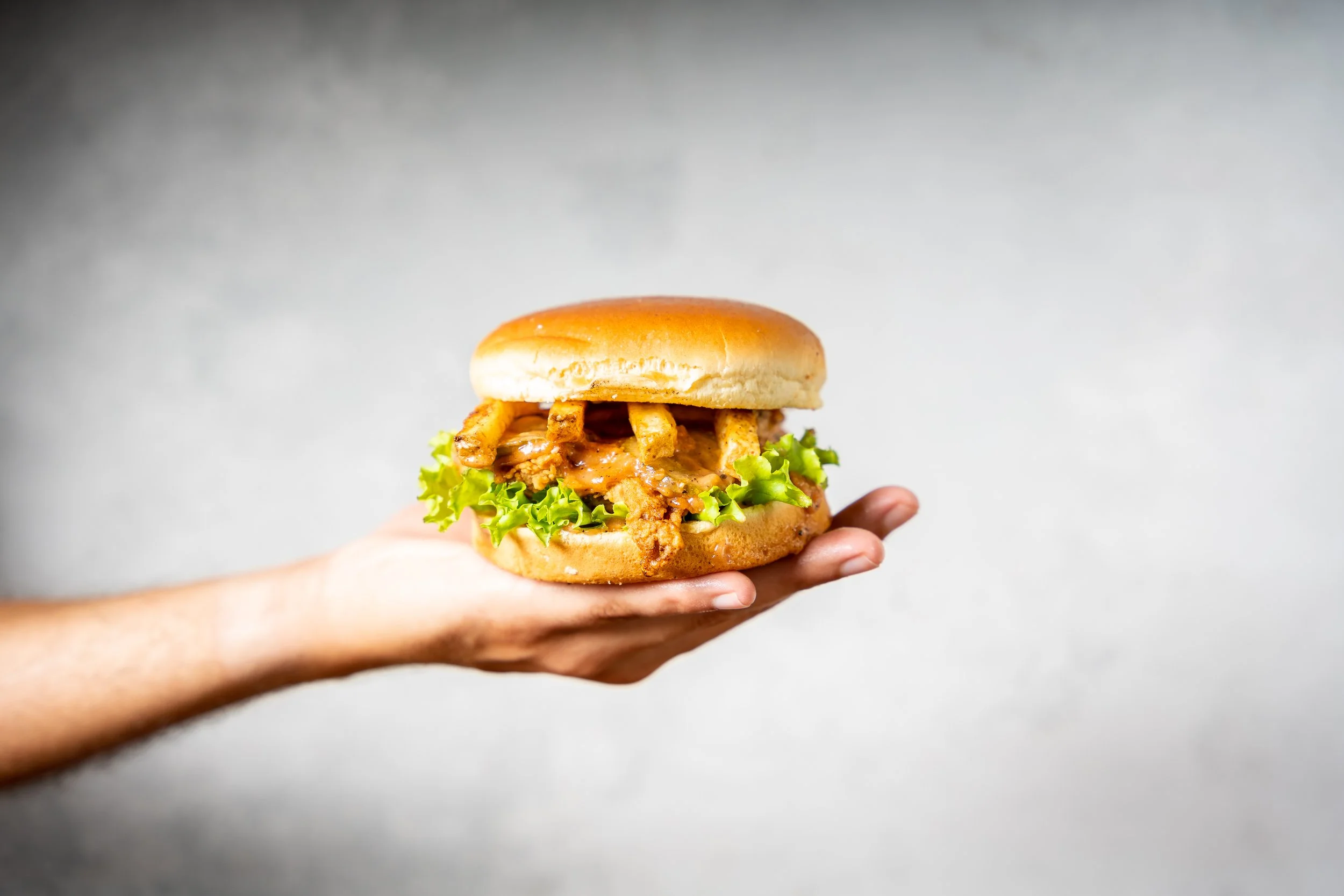 Hand holding a burger with fried chicken, lettuce, and bun against a plain background.
