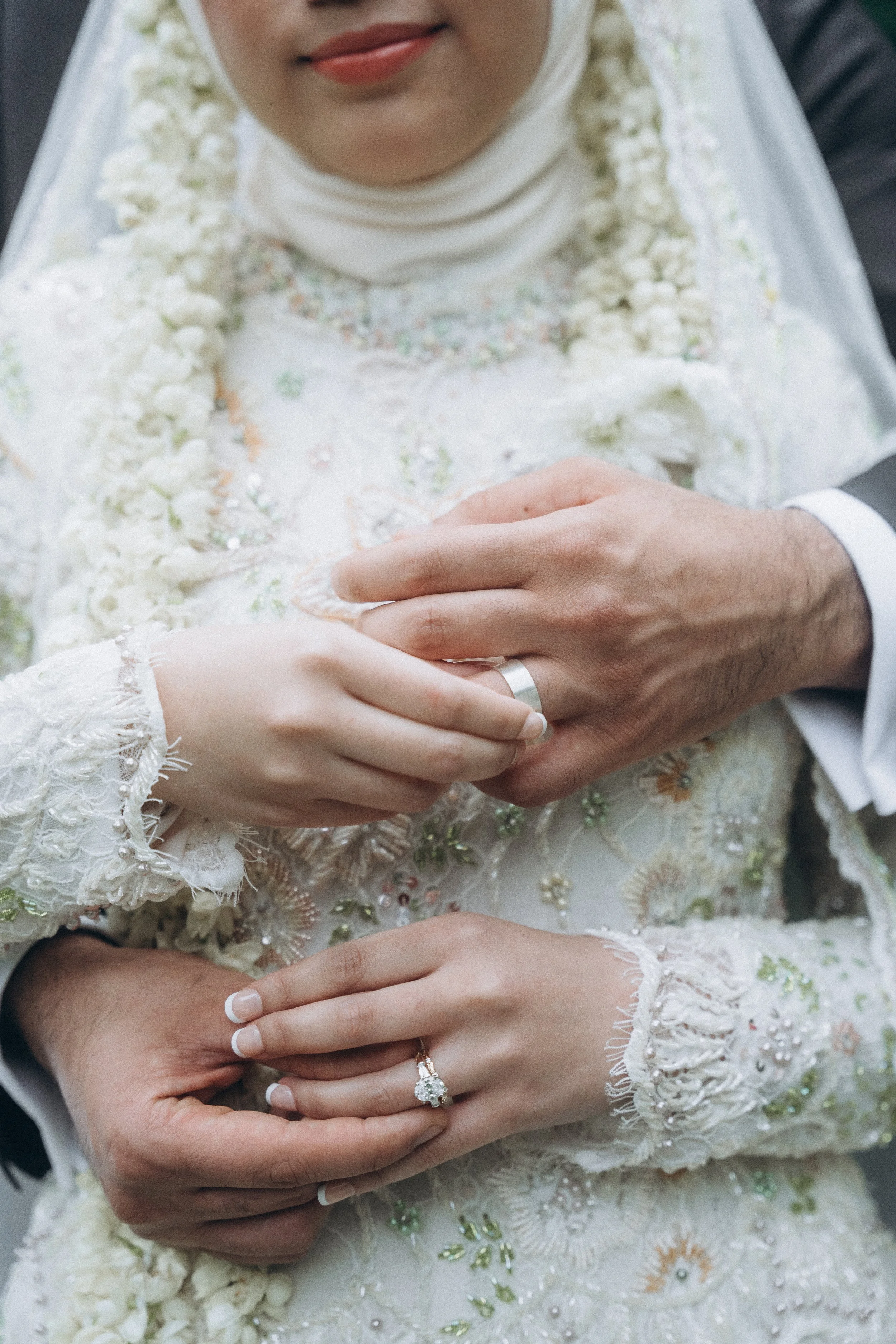 Close-up of a bride and groom holding hands during a wedding ceremony, with wedding rings visible on their fingers and the bride wearing a detailed, embroidered dress.