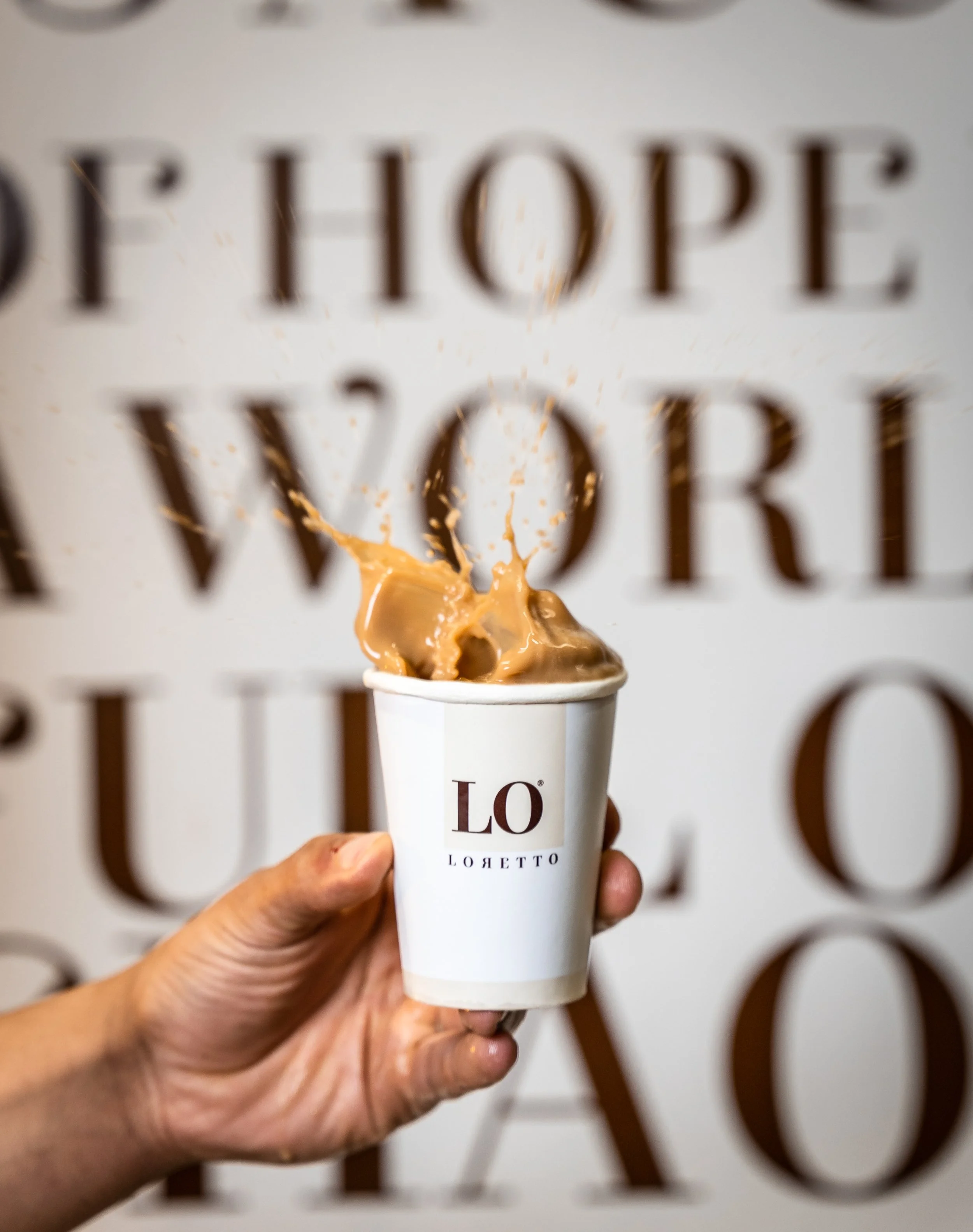 A hand is holding a white cup with the LO logo, with splash of coffee or tea above it, against a background with large brown letters.