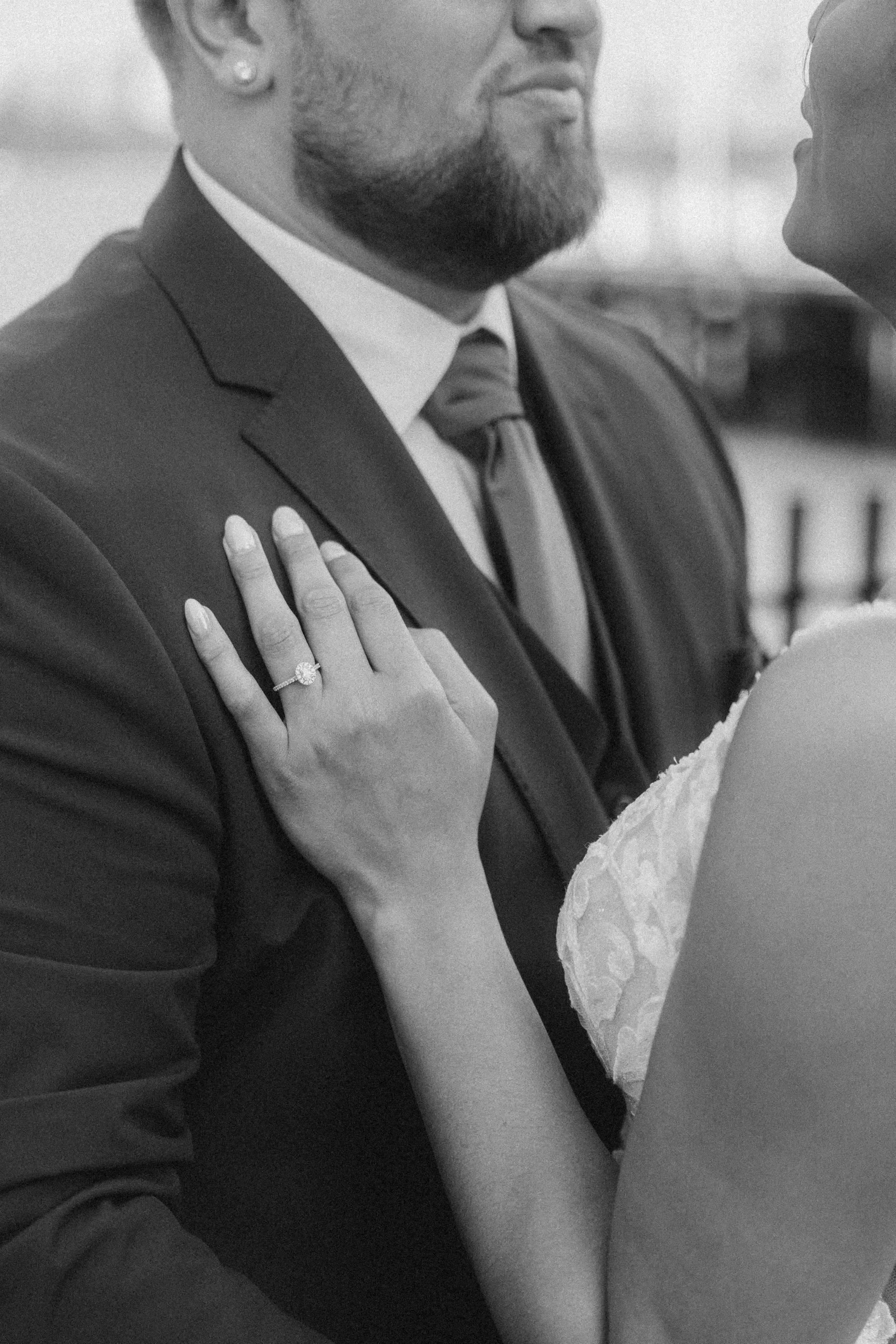 A close-up black-and-white photo of a couple during their wedding, the woman showing her engagement ring on her finger while holding the man's shoulder during their first dance.