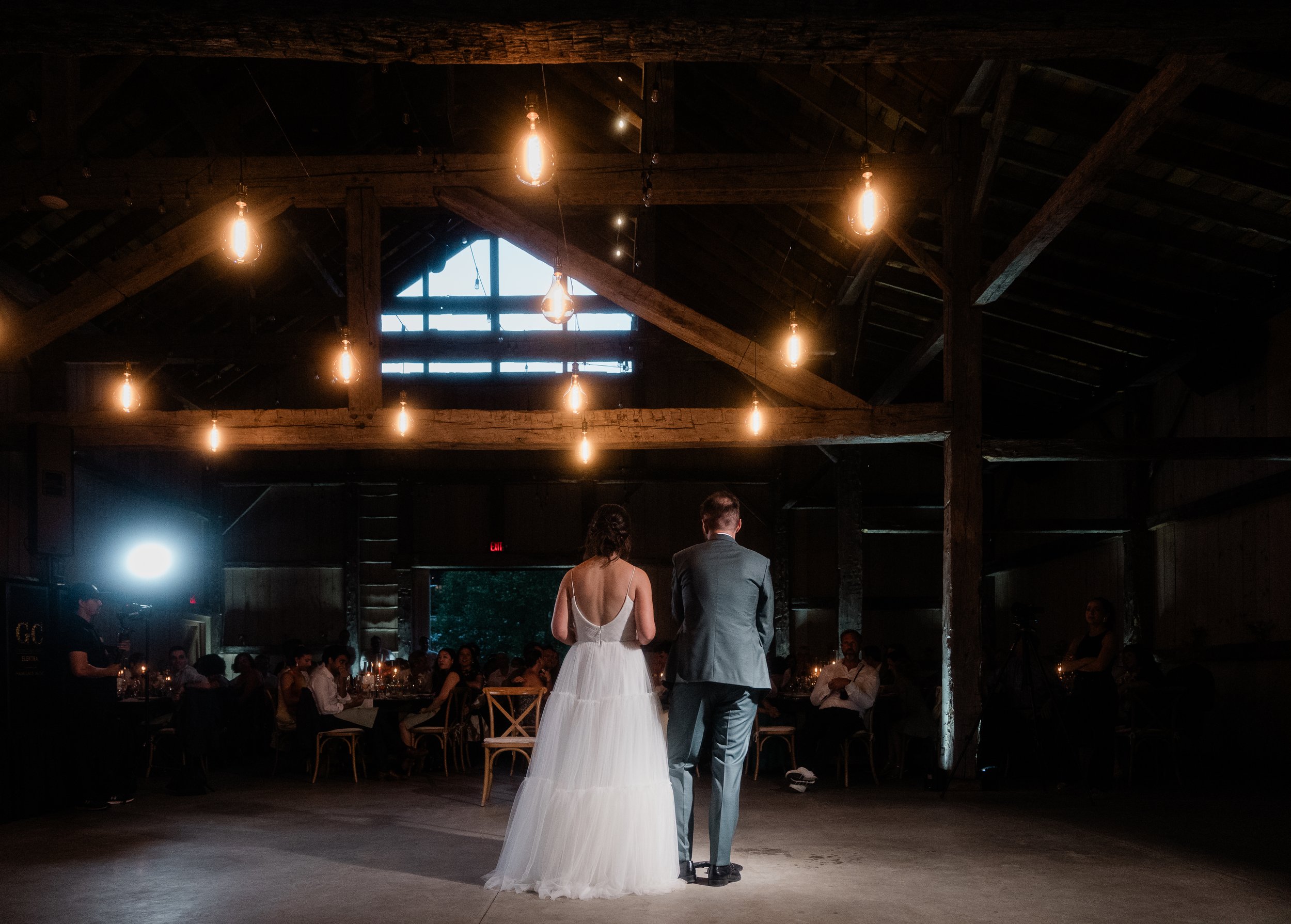 Bride and groom dancing at their wedding reception in a rustic barn with hanging Edison bulbs and surrounded by seated guests.