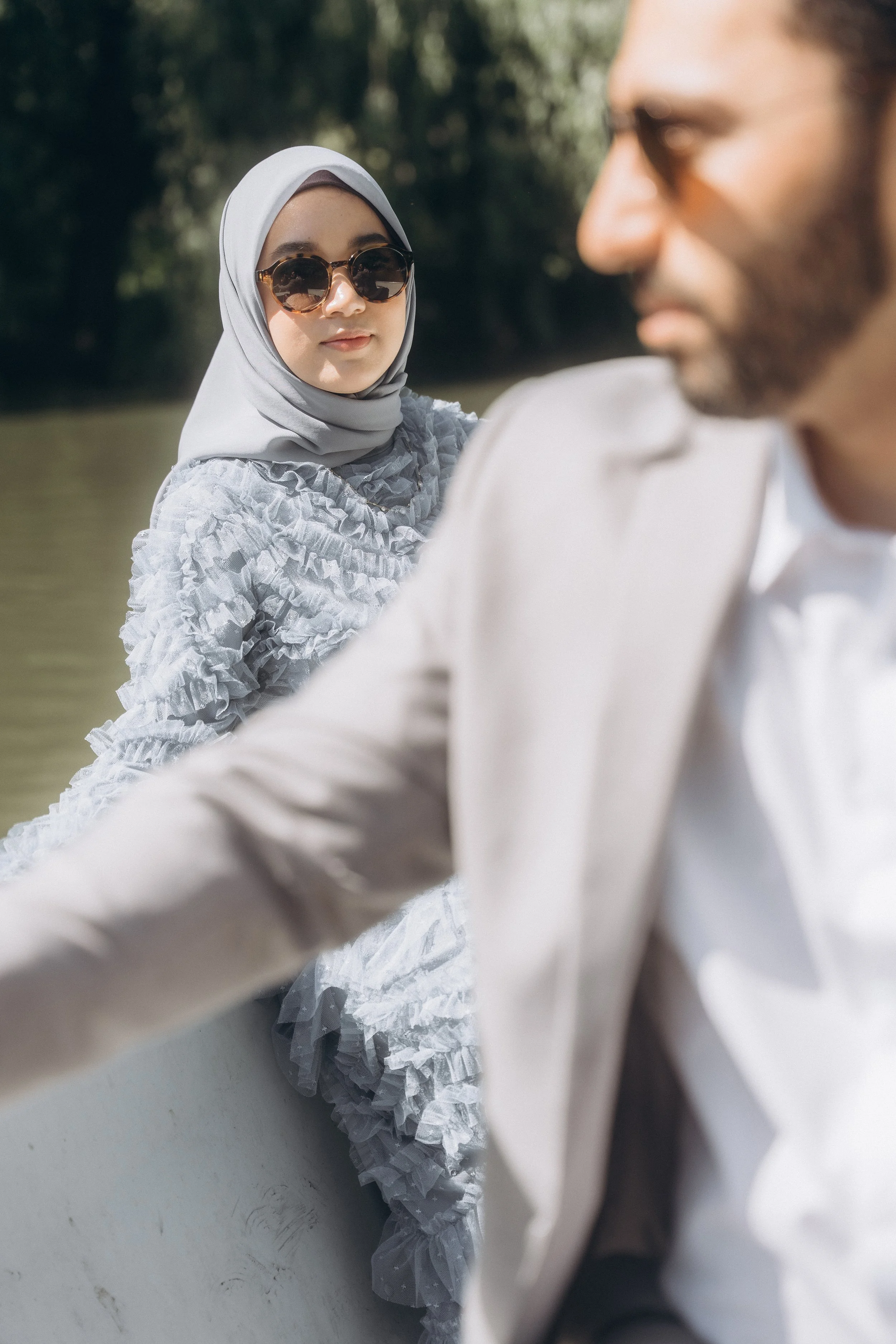 A woman wearing sunglasses and a hijab sitting in a boat on a lake in Central Park NYC, with a man in a suit in the foreground.