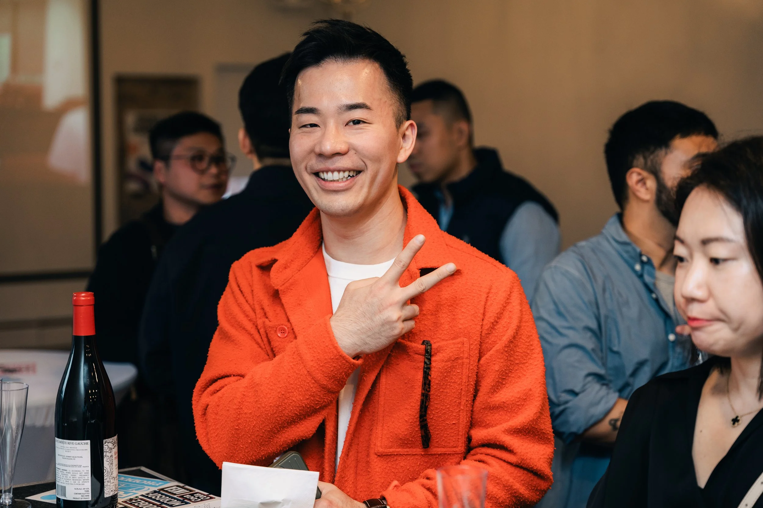 A man in an orange jacket smiling and making a peace sign at a social gathering.