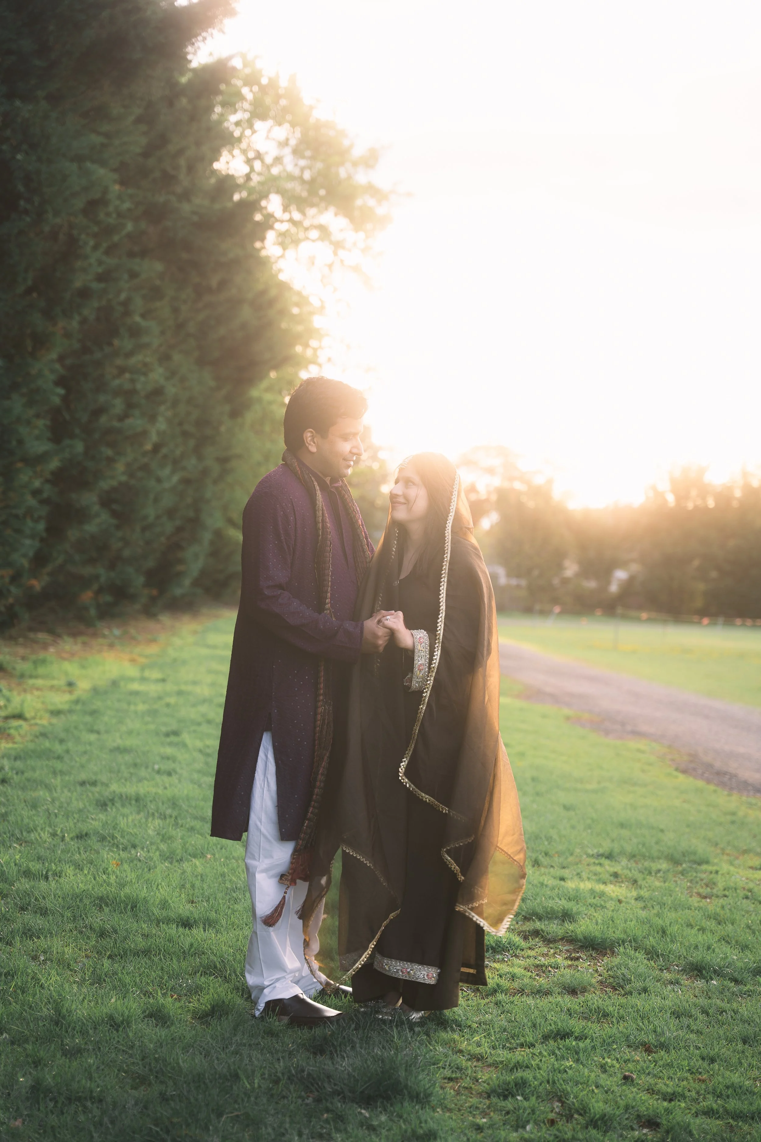 A couple dressed in traditional Indian attire, holding hands and looking at each other in a park during sunset.