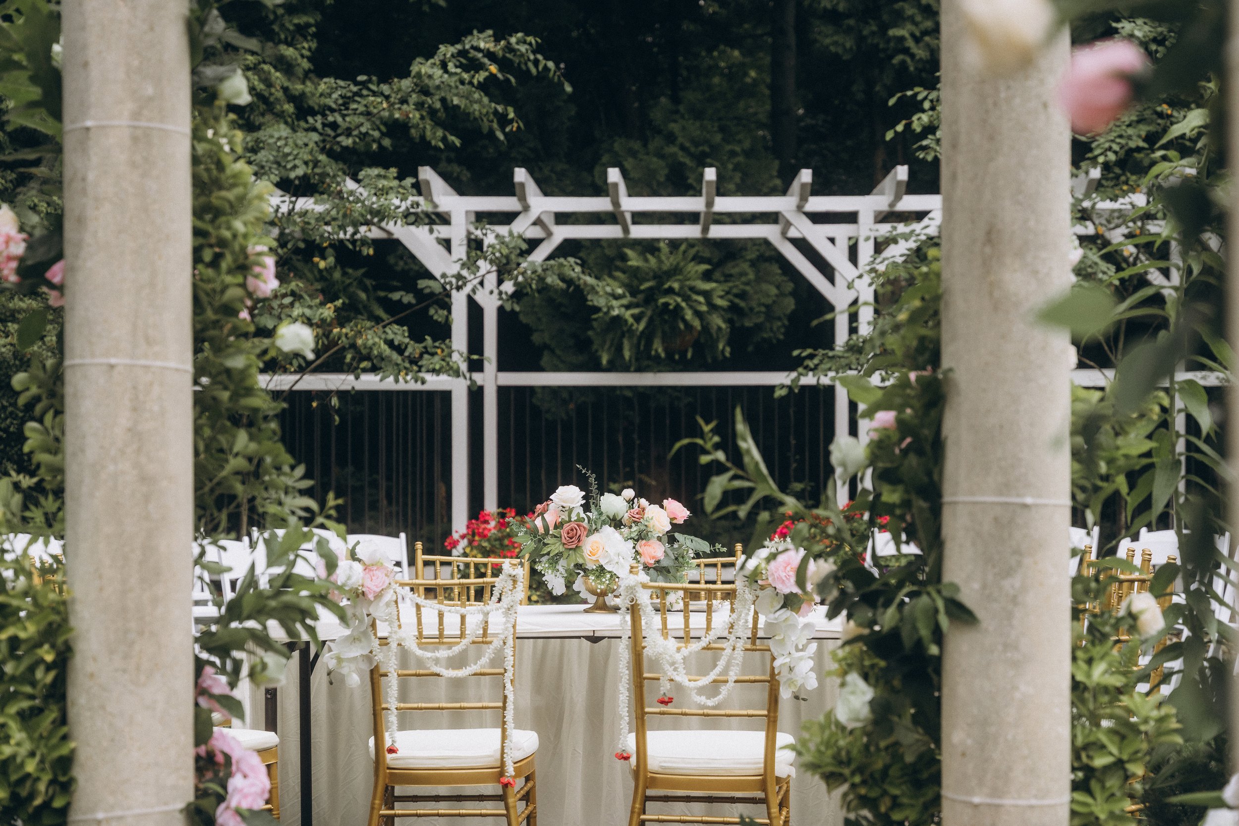 Elegant outdoor wedding table setup with a round table, gold chairs decorated with floral garlands, and a centerpiece of white, pink, and blush flowers on a white tablecloth, framed by stone columns and surrounded by greenery and flowering plants.