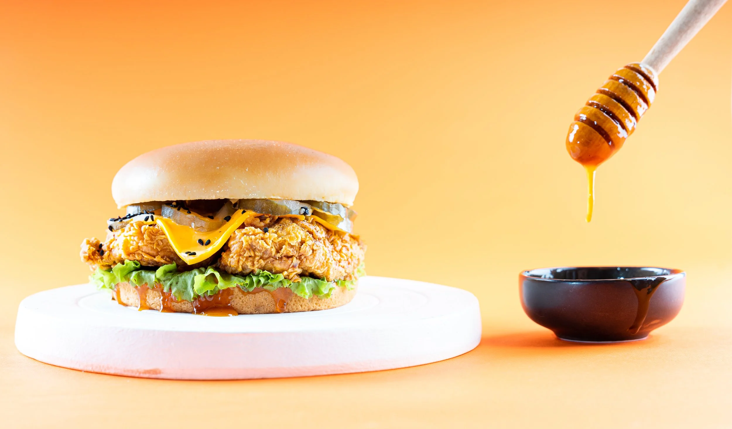 Fried chicken sandwich with pickles, cheese, lettuce, tomato, and bun on a white stand, honey dripping from a honey dipper into a small black dish on an orange background.