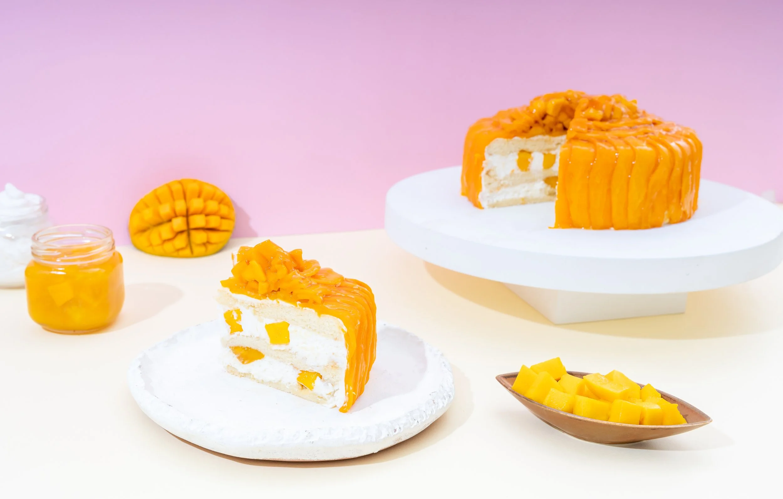 A slice of mango mousse cake with mango pieces on top, on a white plate, with a larger mango cake in the background, and mango pieces in a brown dish, set against a pink and white background.