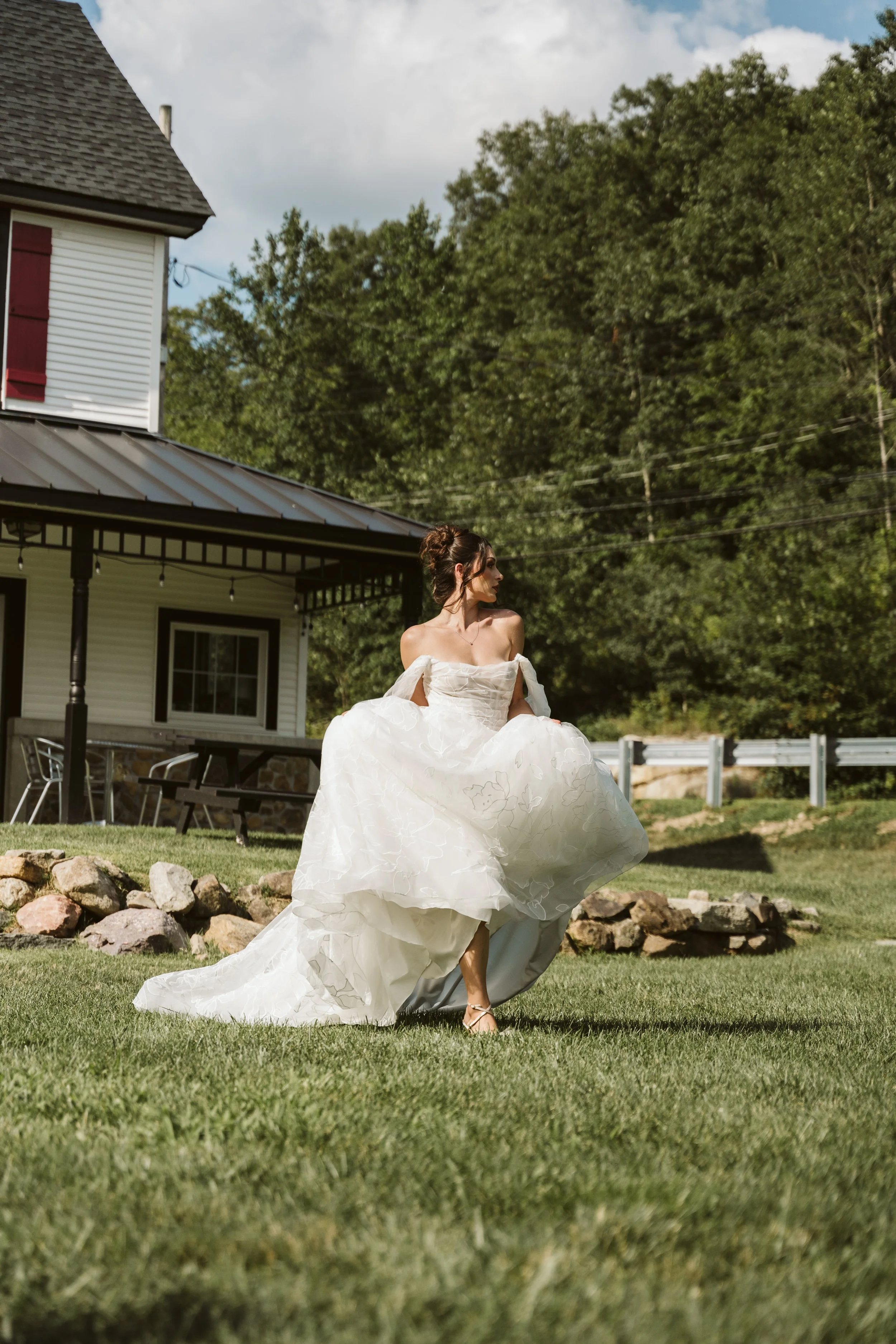 Bride in a white wedding dress standing on a green lawn in front of a house with a porch, trees, and a blue sky with clouds.