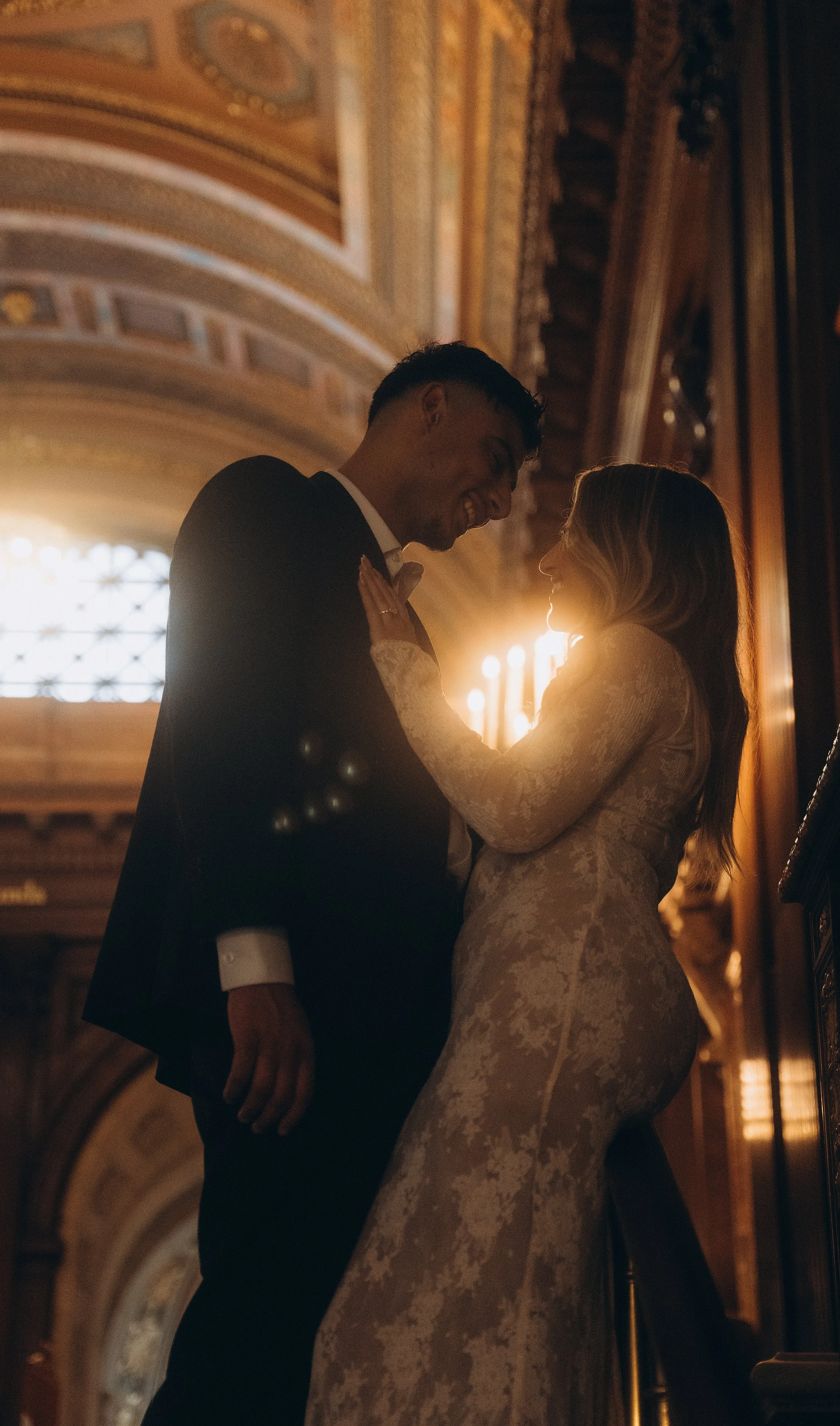 A man and woman sharing an intimate dance in an elegant, ornately decorated hall, backlit by warm lighting.
