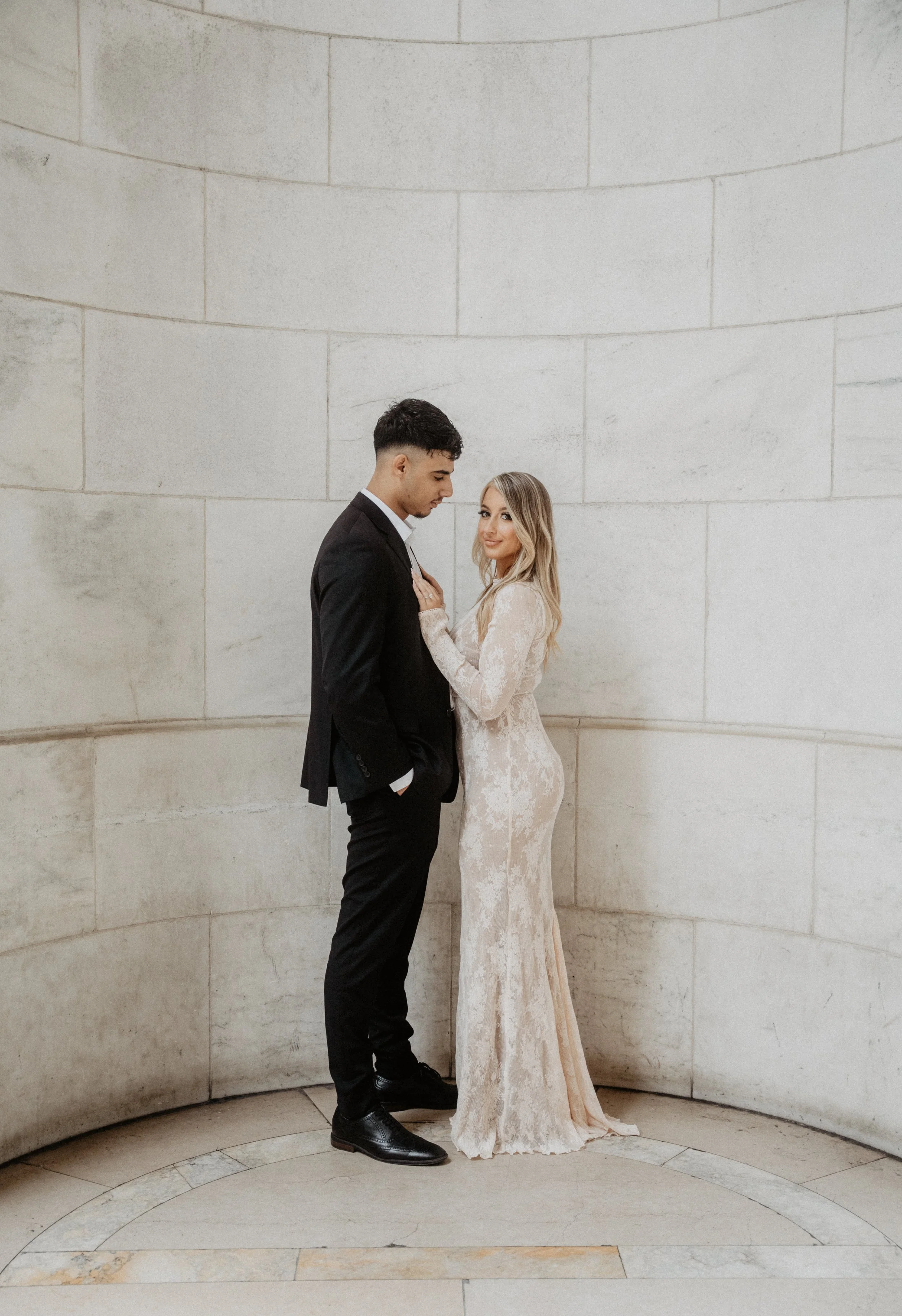 A couple dressed in wedding attire standing close together inside a curved stone wall, with the woman touching the man's chest and smiling at the camera.