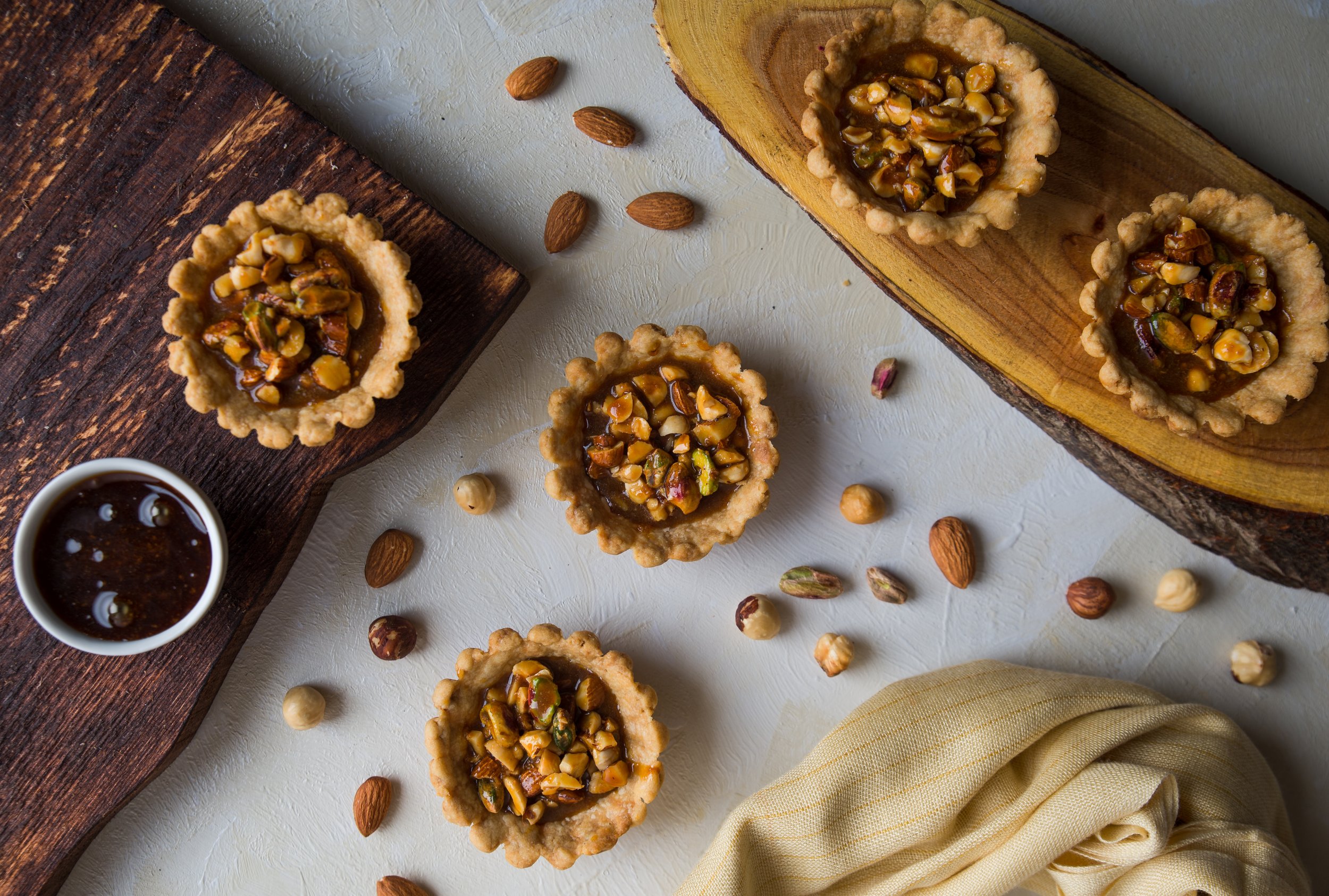 Assorted nut and caramel tartlets on wooden and marble surfaces, with scattered whole nuts and a small bowl of caramel.