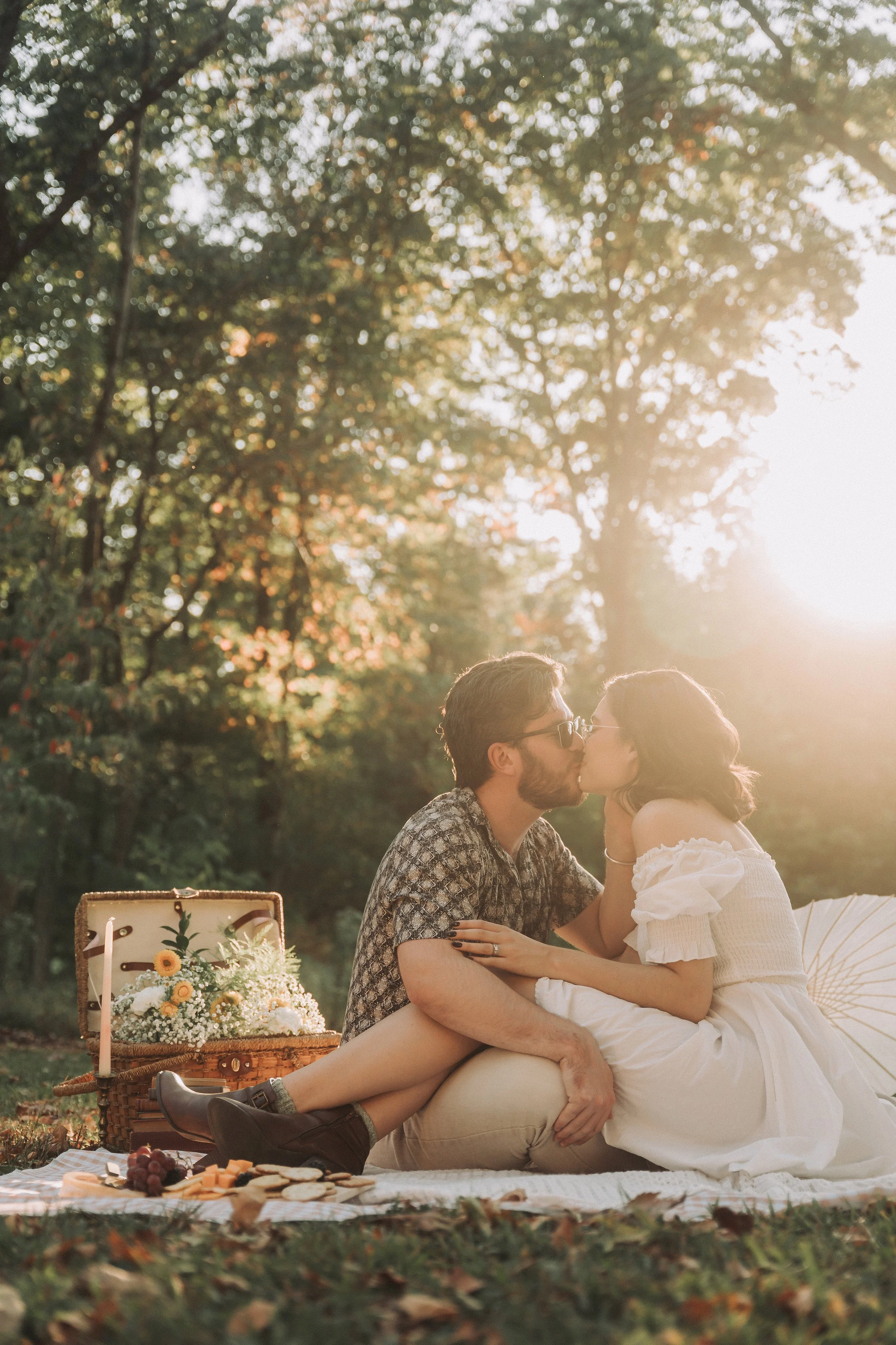 A couple sharing a kiss during a picnic in a park with trees and sunlight in the background.