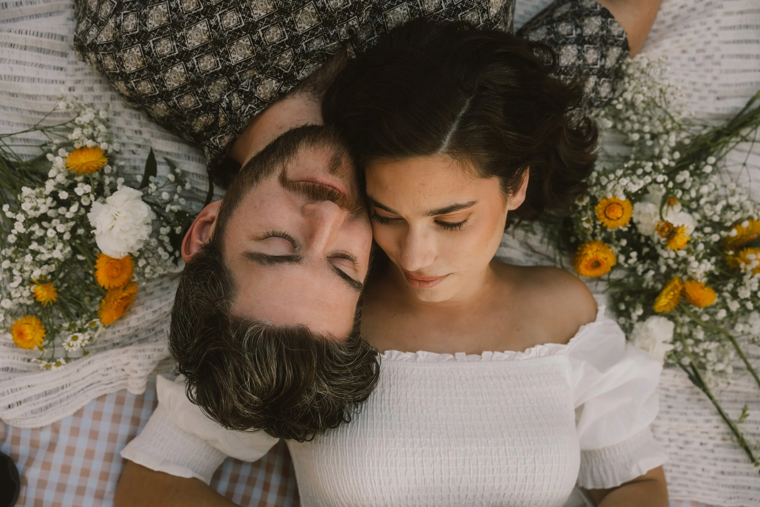 A man and a woman lie on a bed surrounded by small white and yellow flowers with their heads touching, eyes closed, creating a peaceful and intimate scene.