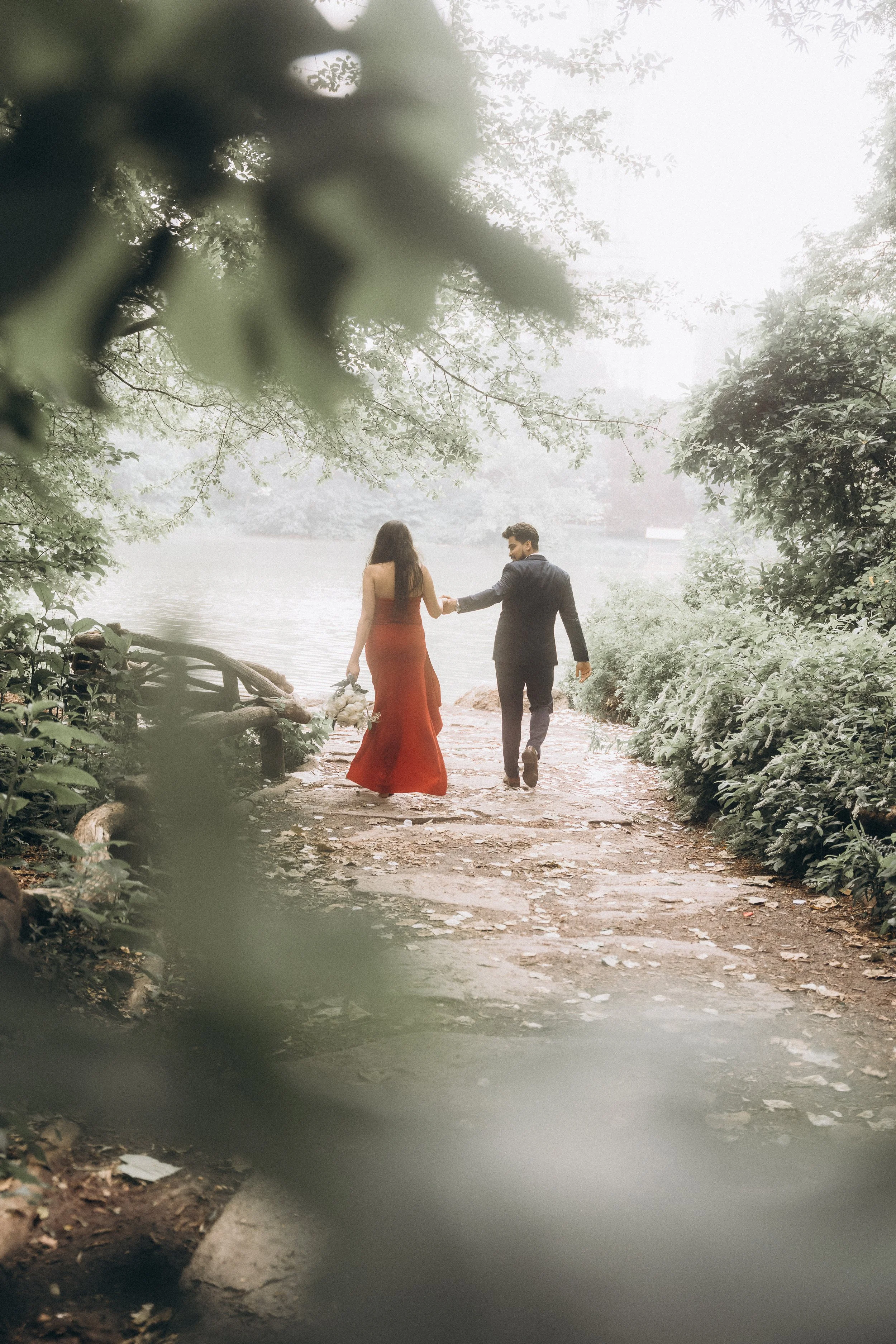 A couple dressed in elegant attire walking hand-in-hand along a foggy lakeside path surrounded by lush greenery in Central Park NYC, with the woman holding a bouquet of flowers.