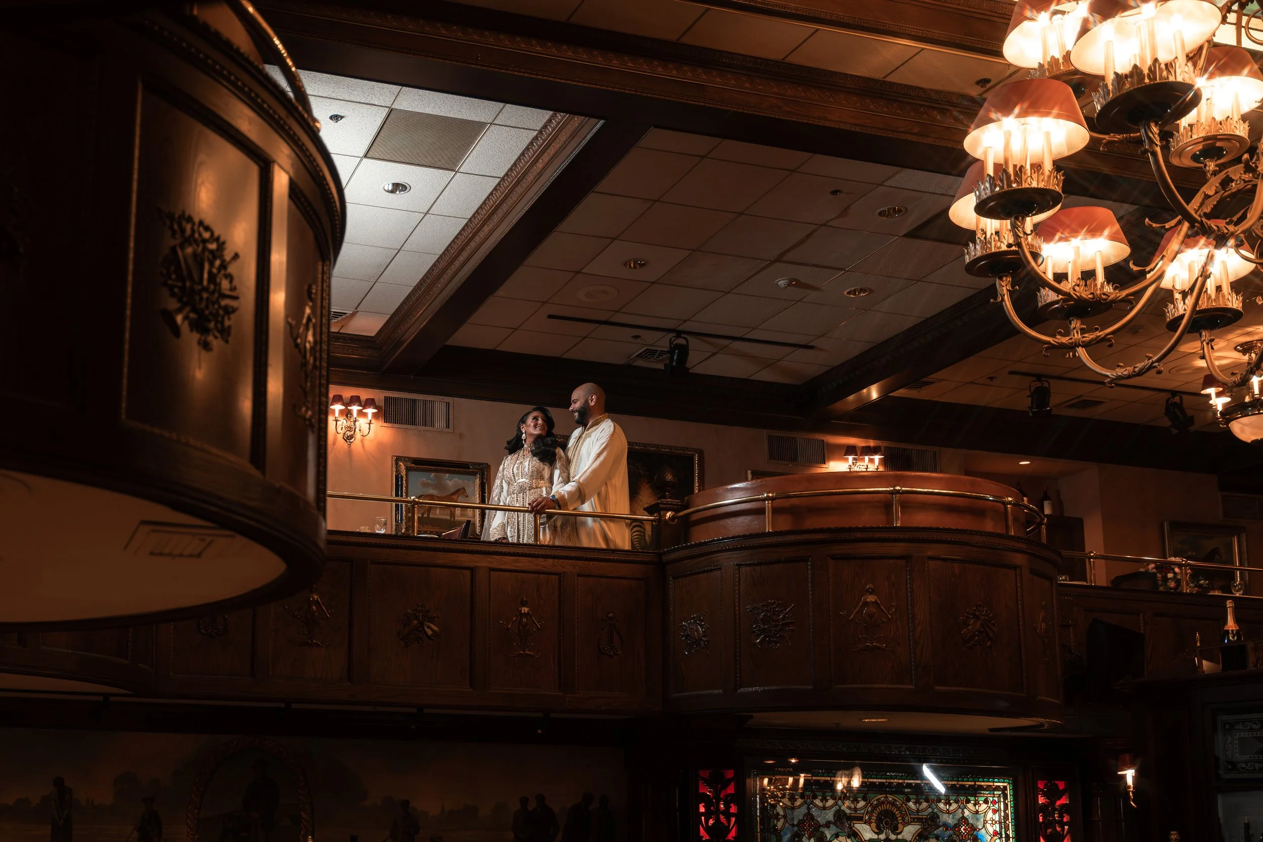 A couple dressed in traditional Indian attire standing on an upper level balcony of a richly decorated banquet hall, surrounded by ornate wood paneling and warm lighting from chandeliers.