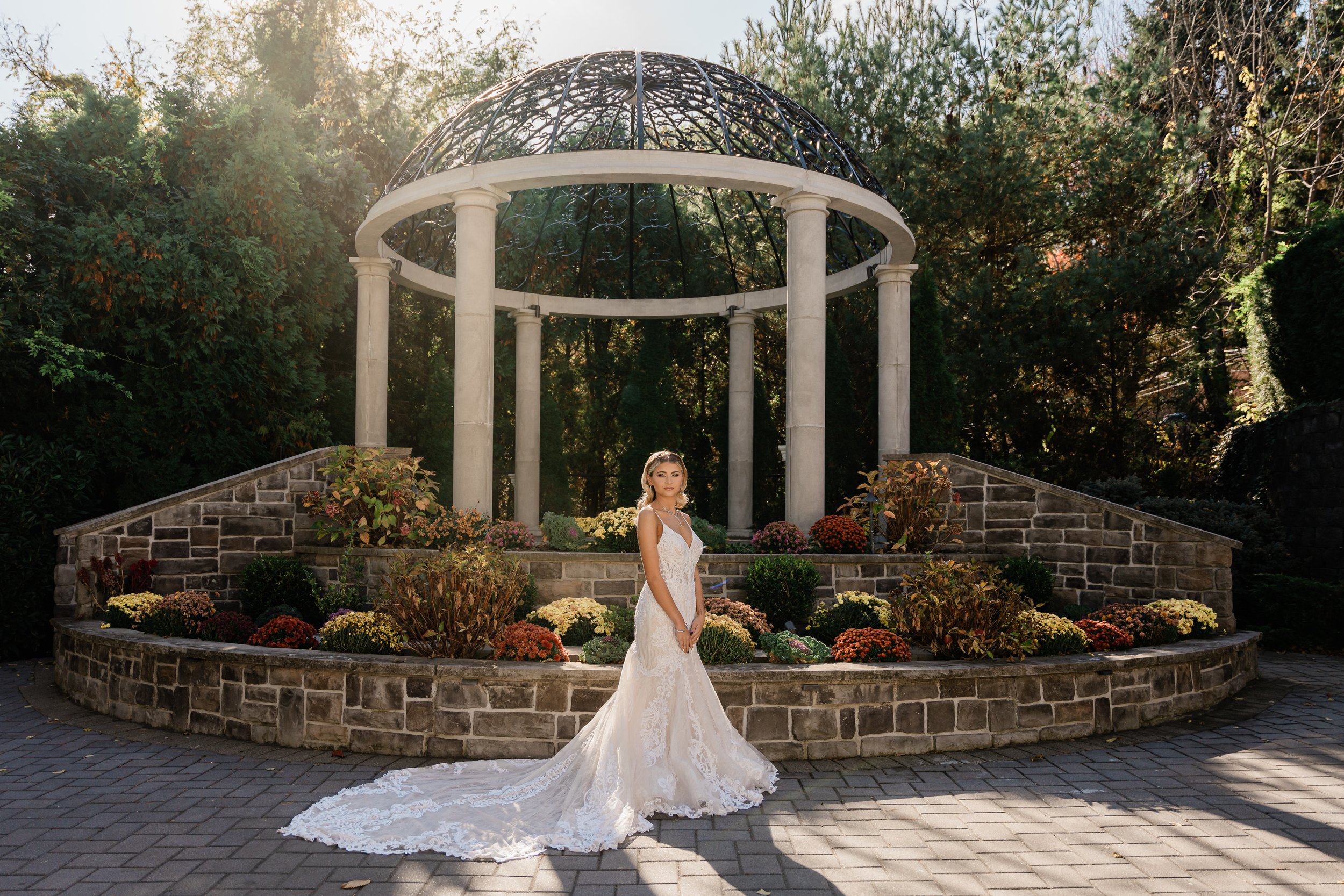 A bride in a white wedding gown standing outdoors in front of a landscaped stone and concrete gazebo with columns, surrounded by colorful flowers and greenery, during daylight.