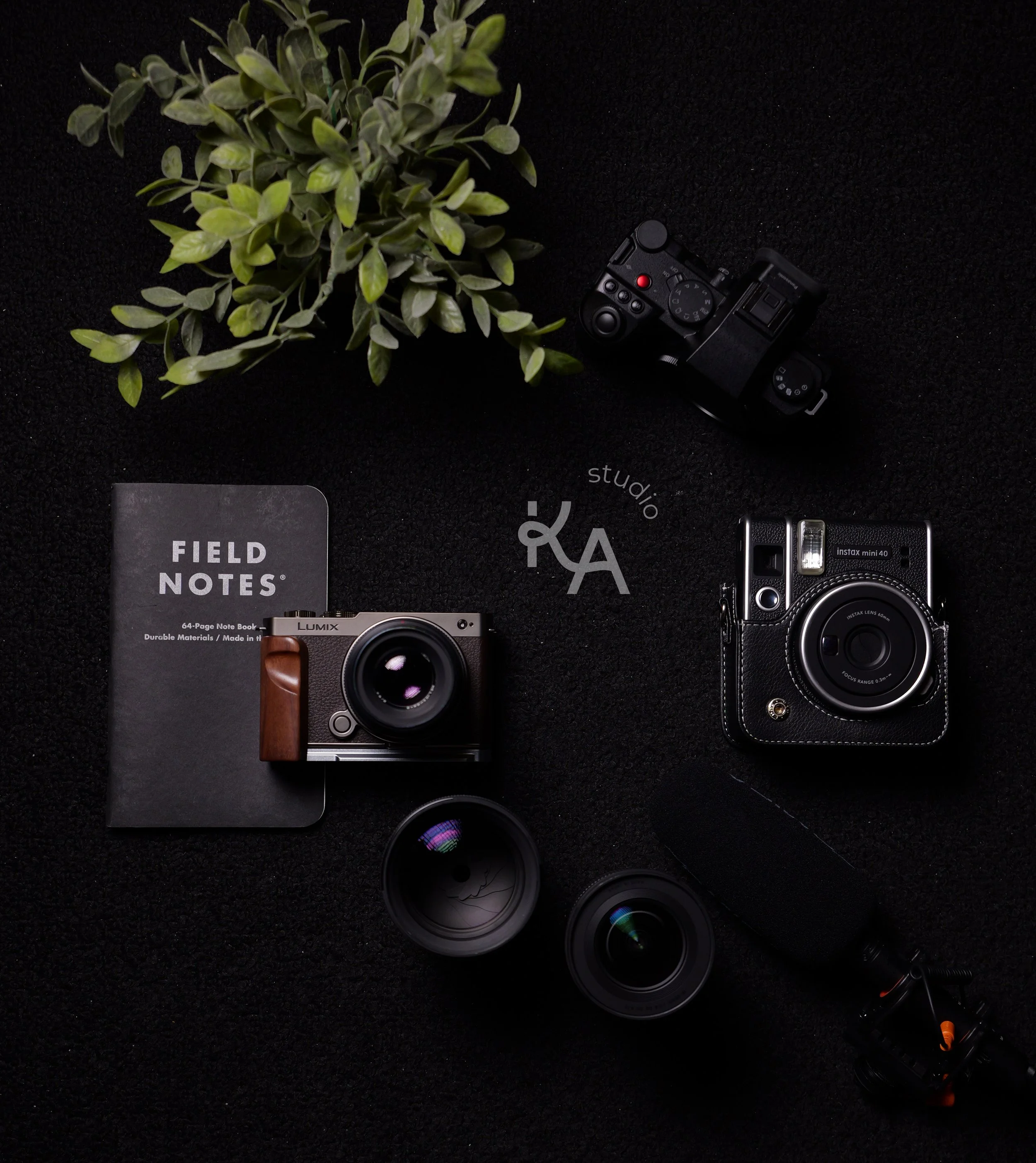 Photography equipment laid out on a black surface, including two cameras, two camera lenses, a small microphone, a notepad titled 'Field Notes,' and a potted plant.