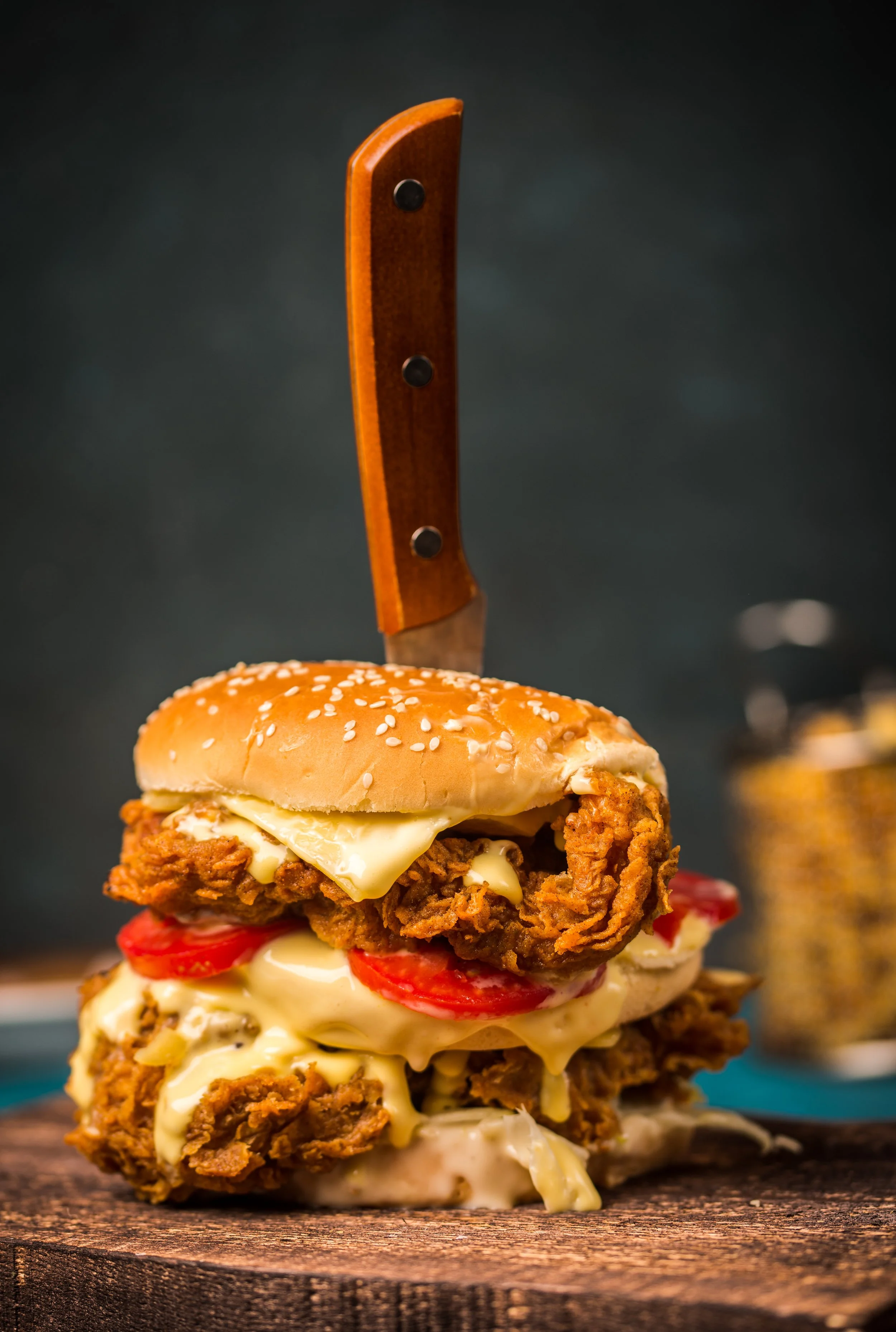 A towering fried chicken sandwich with a cheeseburger layer, tomato slices, and mayonnaise on a wooden surface, with a knife inserted through the top bun