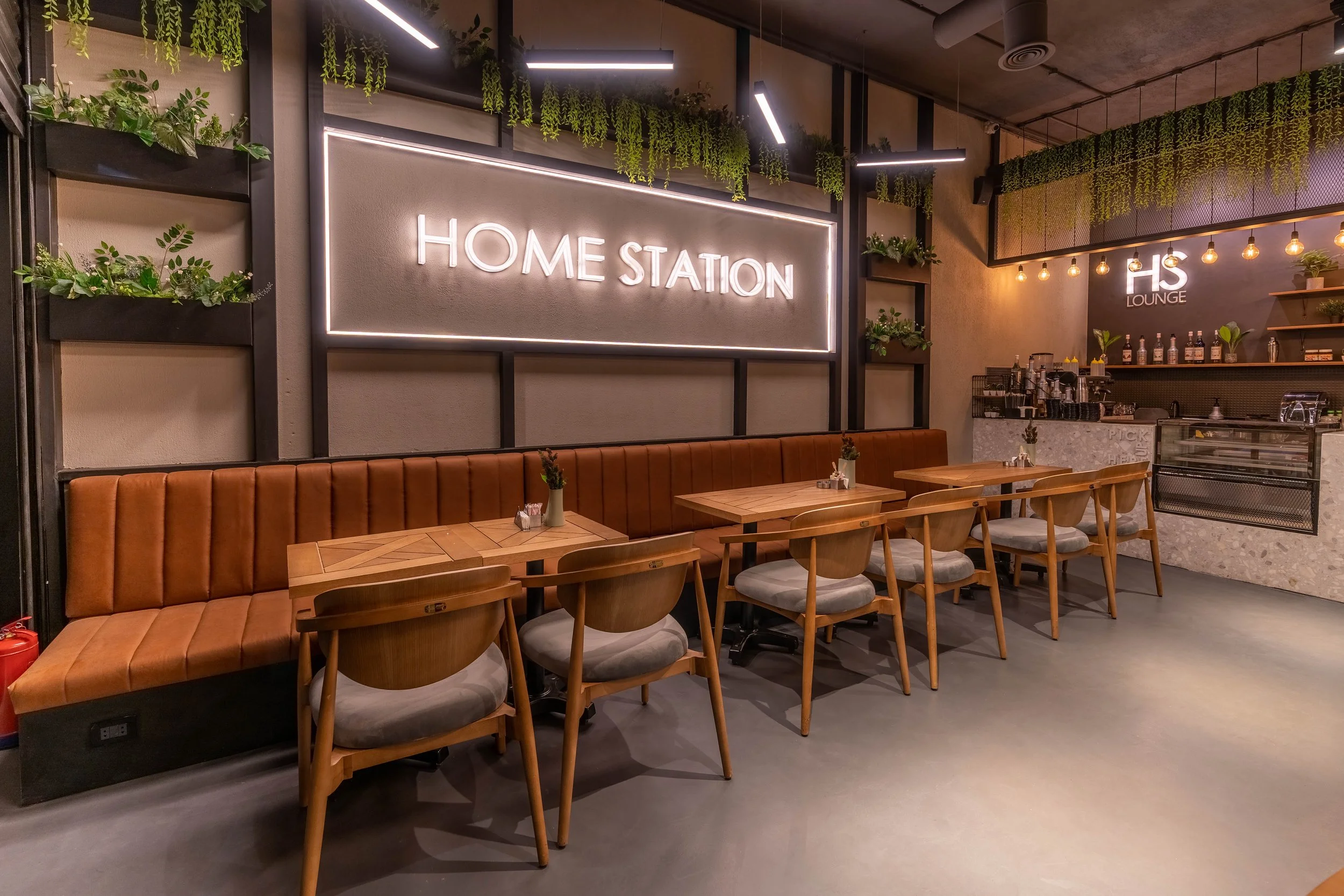 Interior of a modern cafe with a leather banquette, wooden chairs, and tables. Bright illuminated signs display 'HOME STATION' and 'HS Lounge'. Green plants decorate the wall, and the counter area features a coffee machine and bottles.