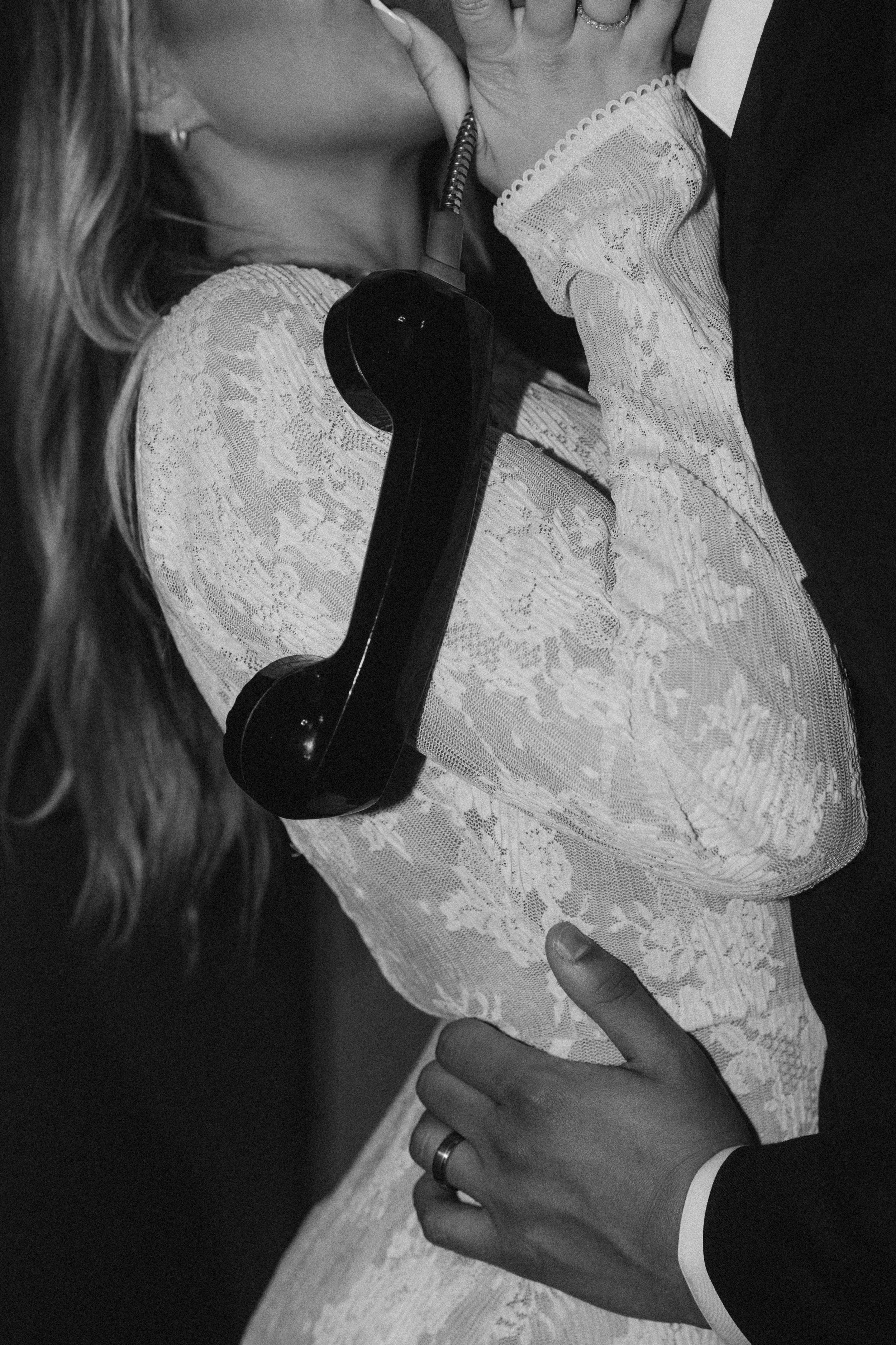 A woman dressed in lace attire leaning in to kiss a man dressed in a tuxedo, while the man holds a vintage black telephone receiver pressed against the woman’s neck.