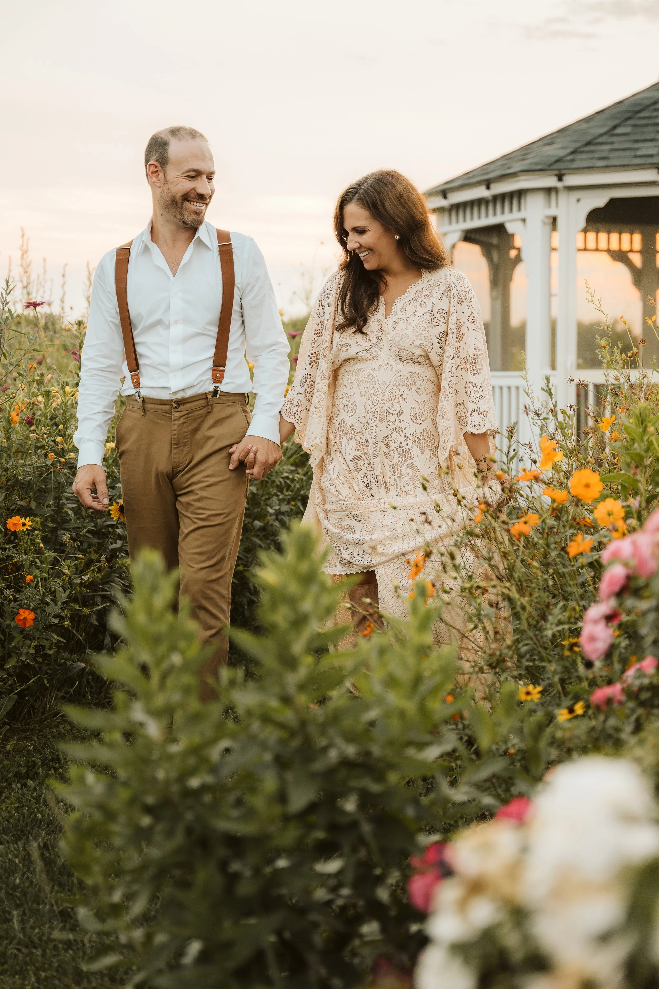 A couple walking hand-in-hand through a garden filled with colorful flowers at sunset, with a gazebo in the background.