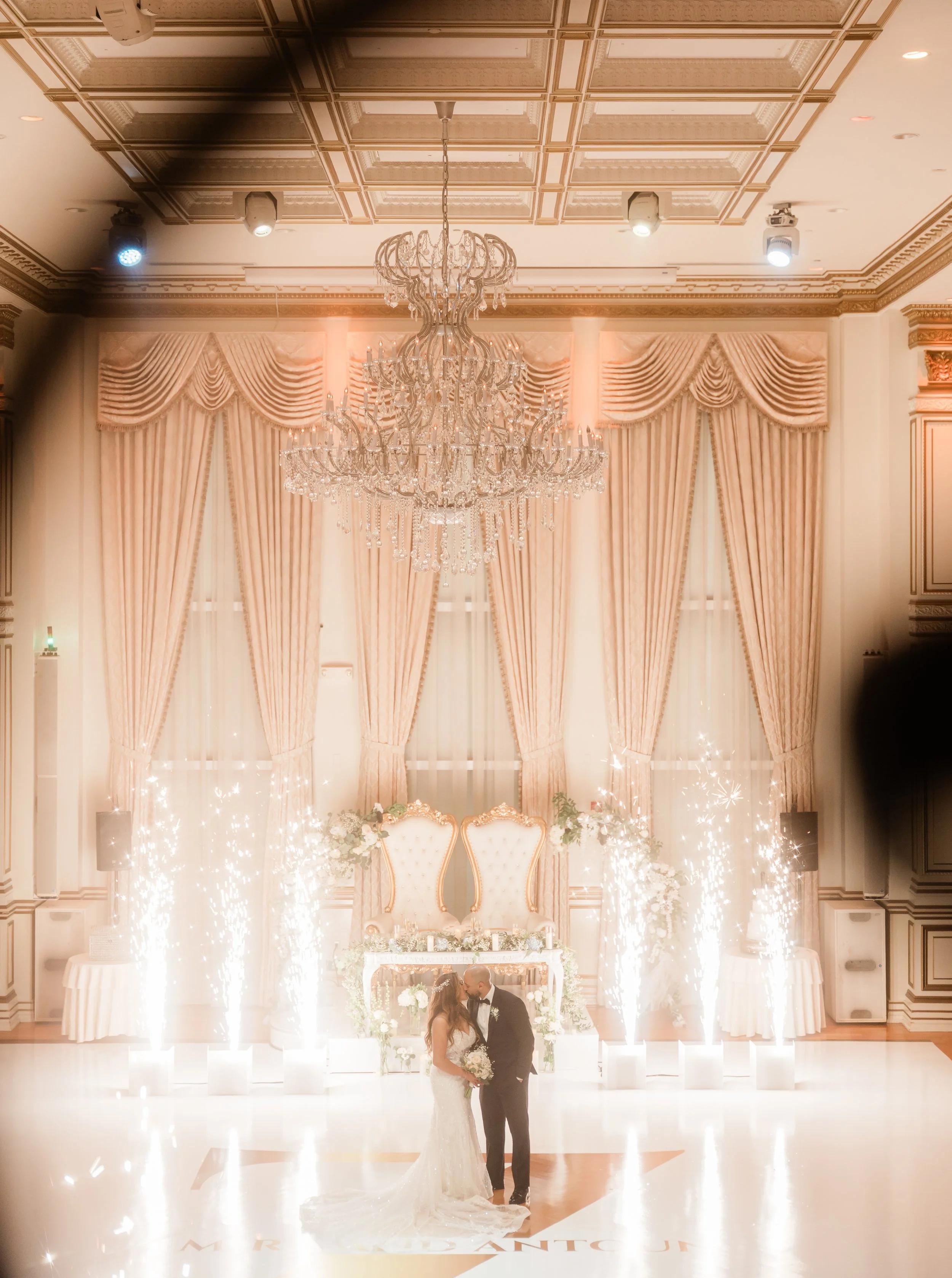 A bride and groom share a kiss during their wedding ceremony in an elegant, decorated ballroom with large windows, pink curtains, a chandelier, and sparklers.