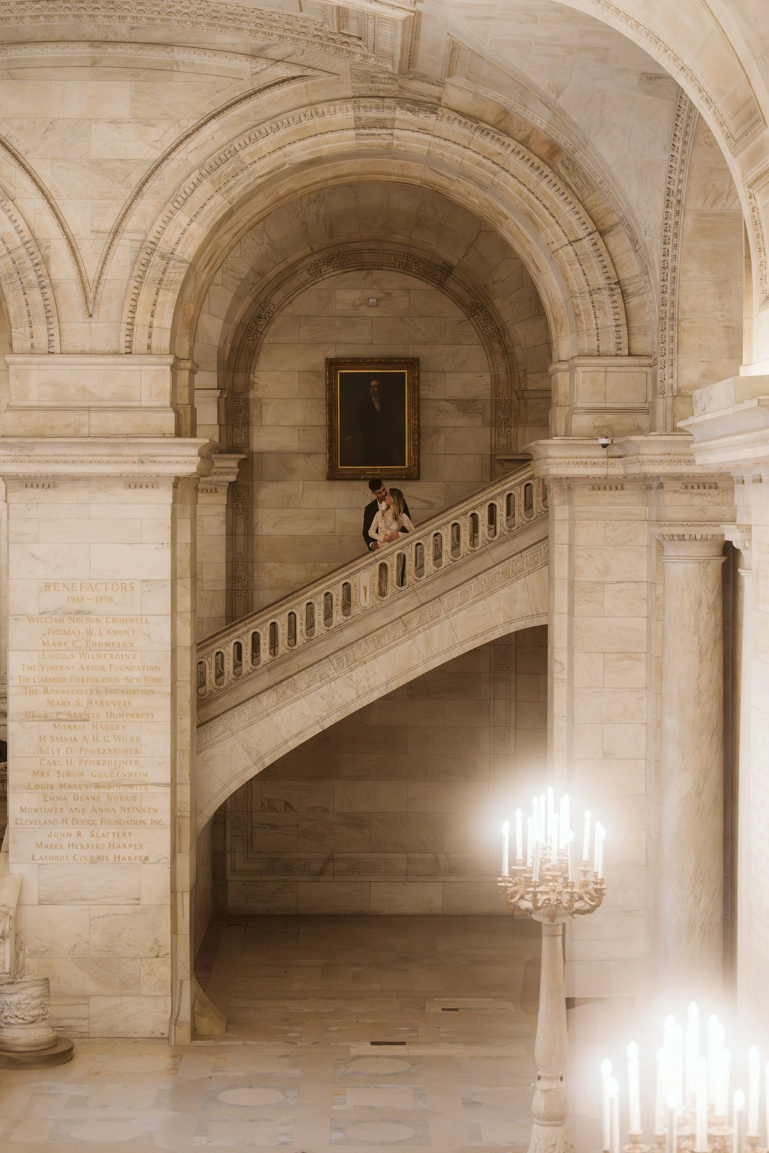 A marble staircase in a historic building with two people, a man and a woman, descending the stairs. The woman leans on the railing, and the man stands close behind her. A portrait hangs on the wall behind them, and there's a large chandelier with li