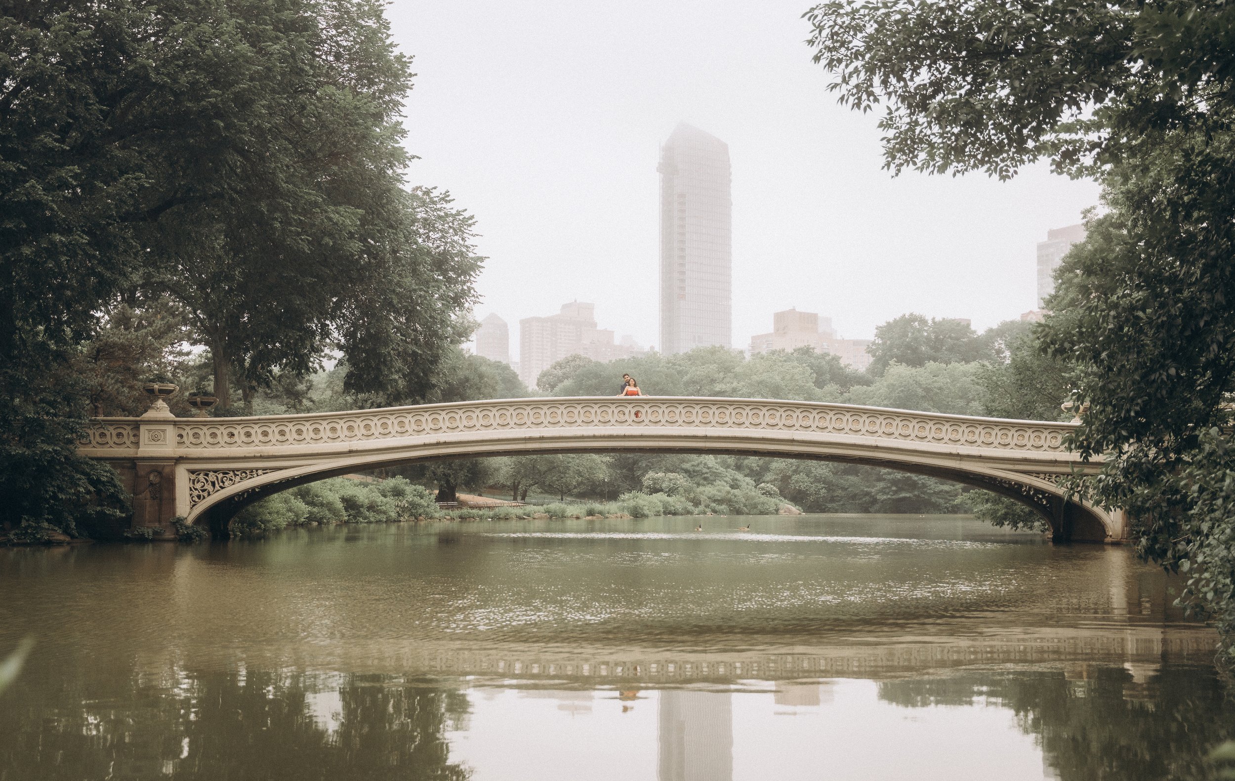 A person stands in the center of a decorative stone bridge (The Bow Bridge) in Central Park NYC over a calm lake in a park with trees and foggy city skyline in the background.