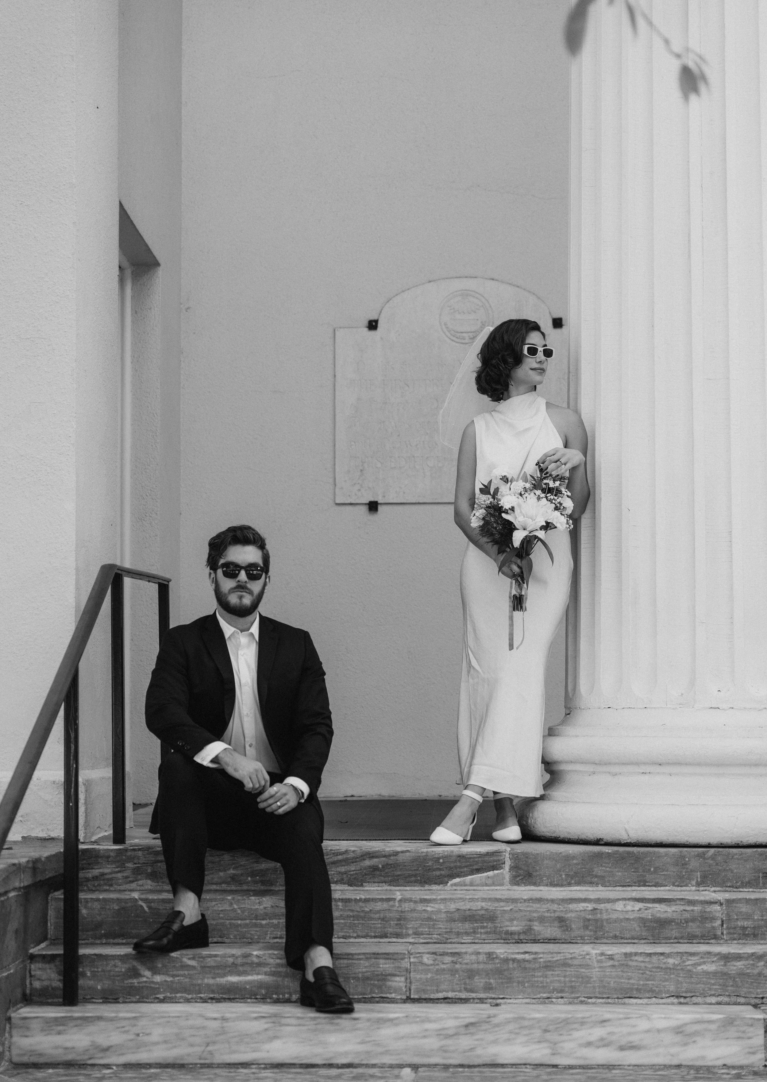 A black and white photo of a woman in a white dress holding a bouquet of flowers, with sunglasses and a veil, standing beside a large classical column. A man in a dark suit and sunglasses is sitting on the steps nearby.