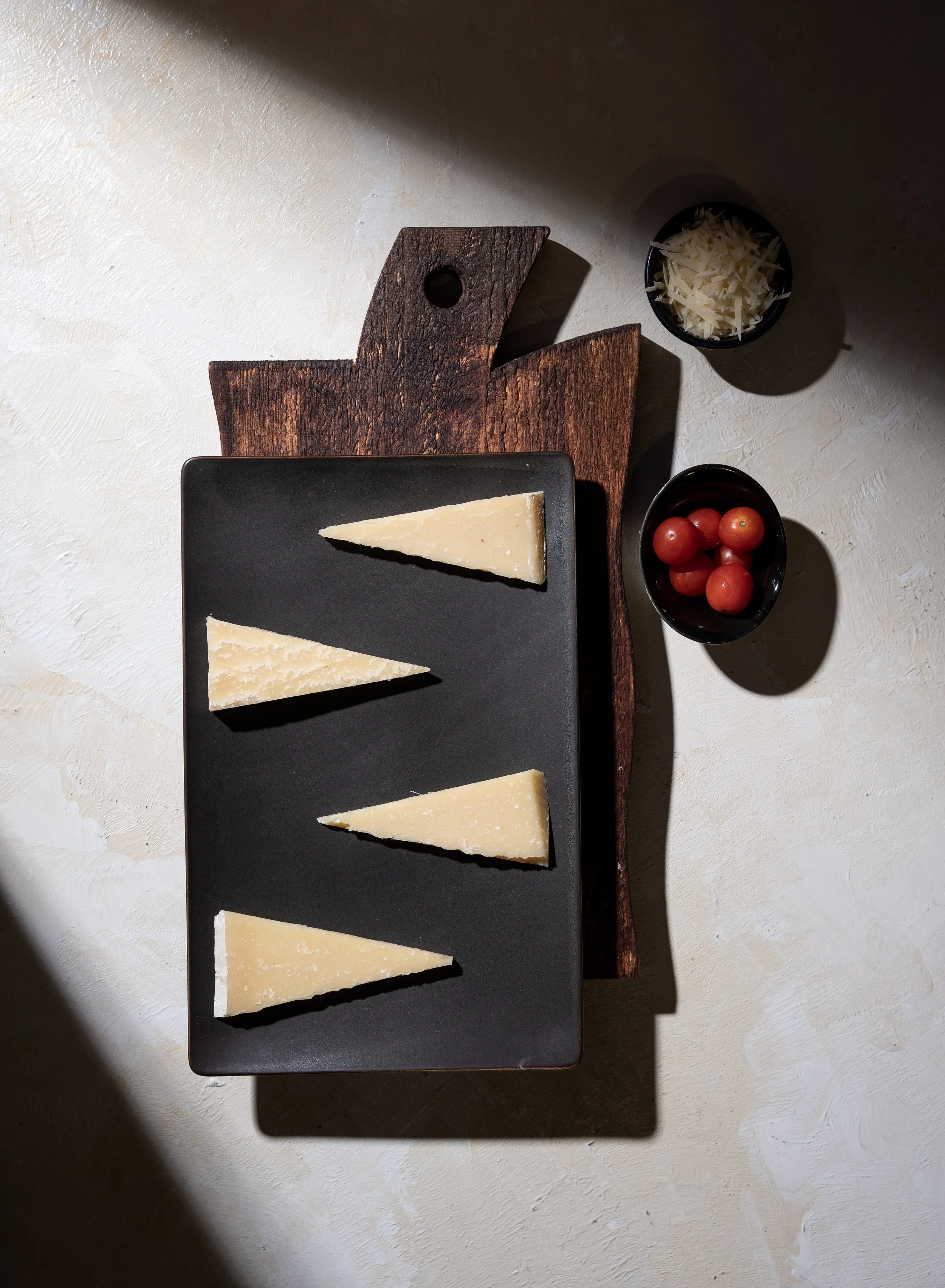 Four wedges of cheese on a black rectangular plate, with cherry tomatoes and shredded cheese in small bowls nearby, on a white textured surface with a rustic wooden board underneath.
