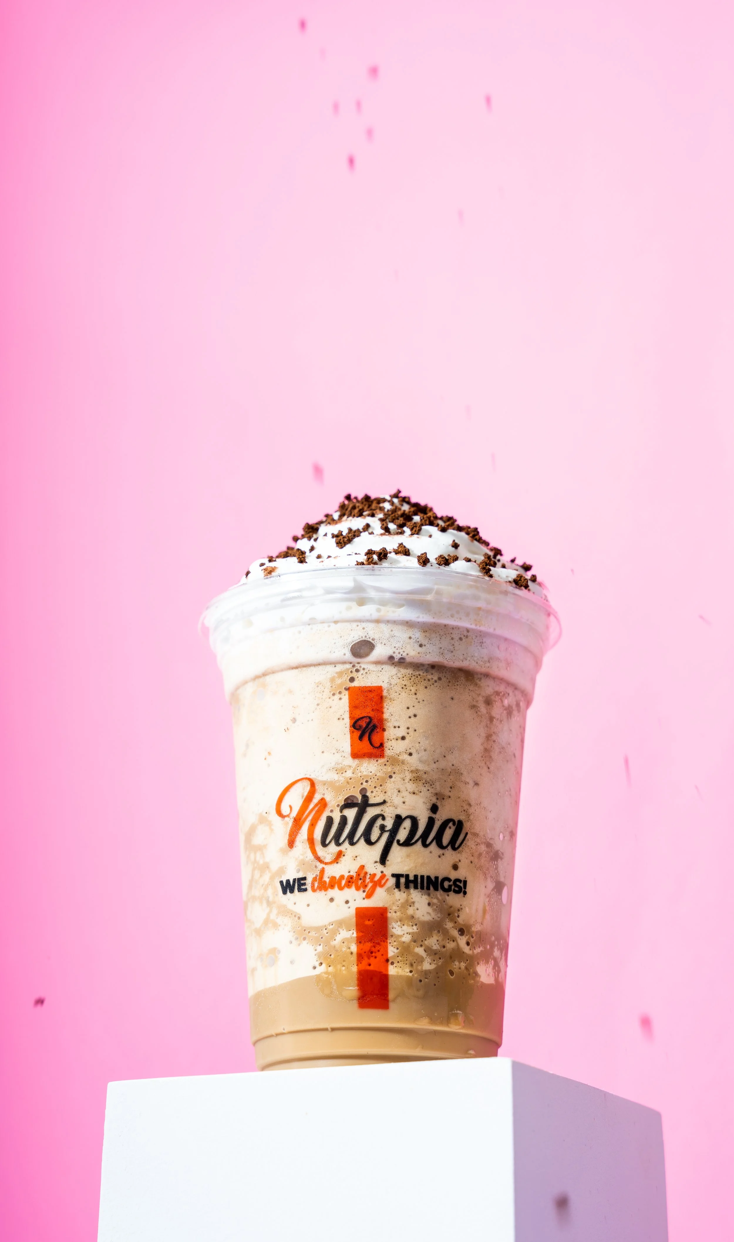 A cold drink topped with whipped cream and chocolate shavings, in a clear cup with Nutopia branding on a white surface against a pink background.