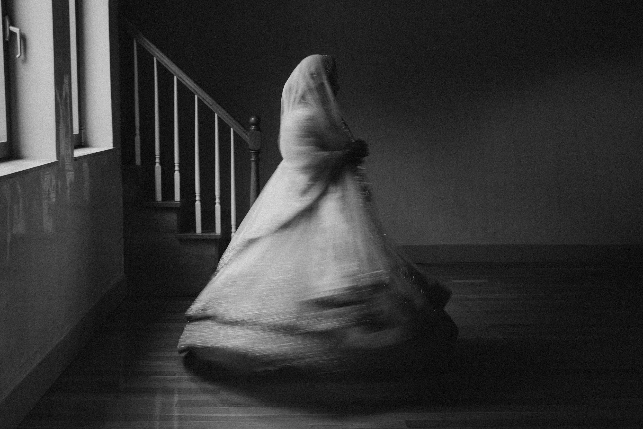 A woman in a full-length gown twirling indoors with stair railing visible on the left, motion blur capturing movement.